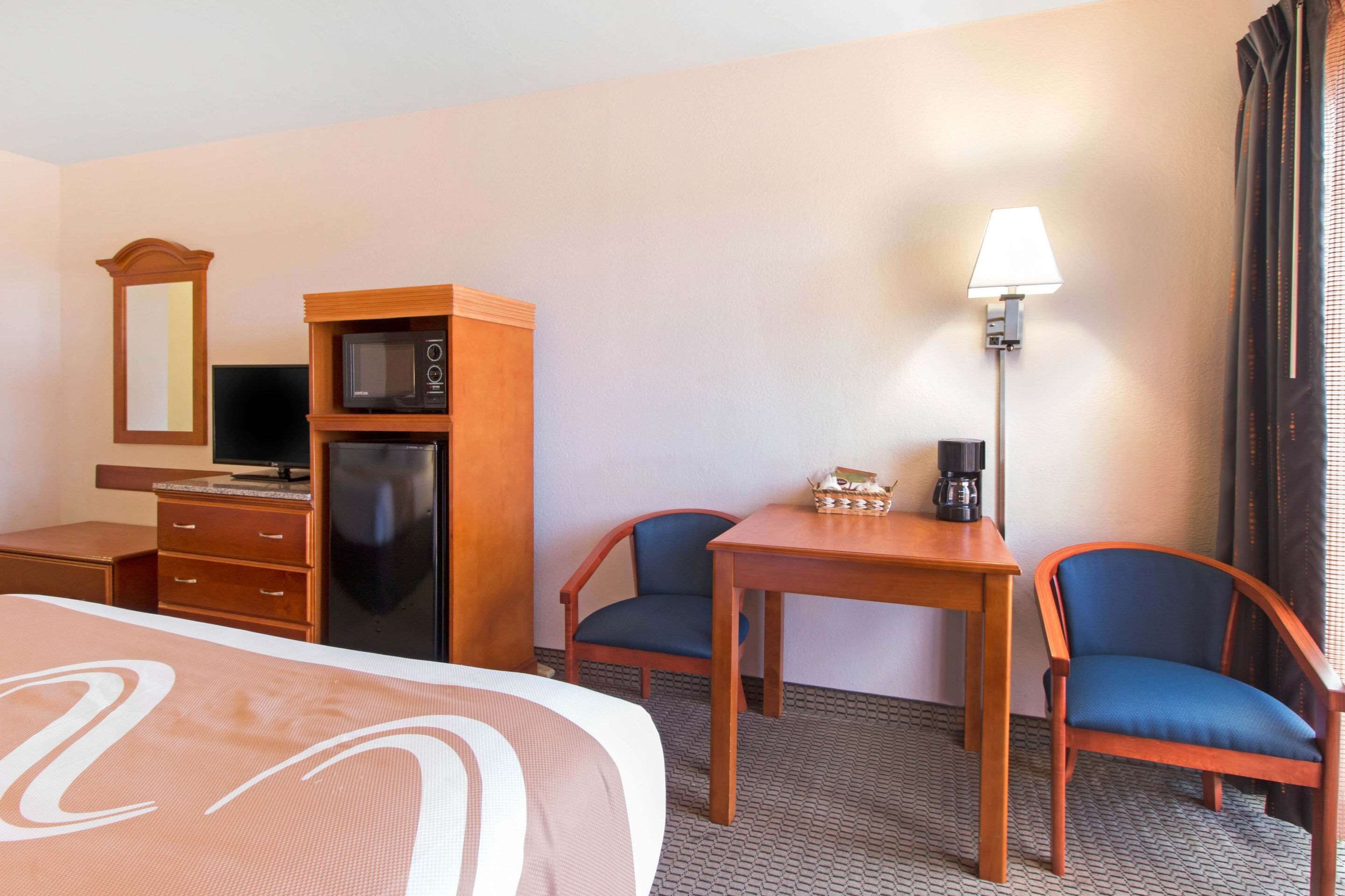 A hotel room with a bed, a desk with a lamp, a microwave, a TV, and two chairs.