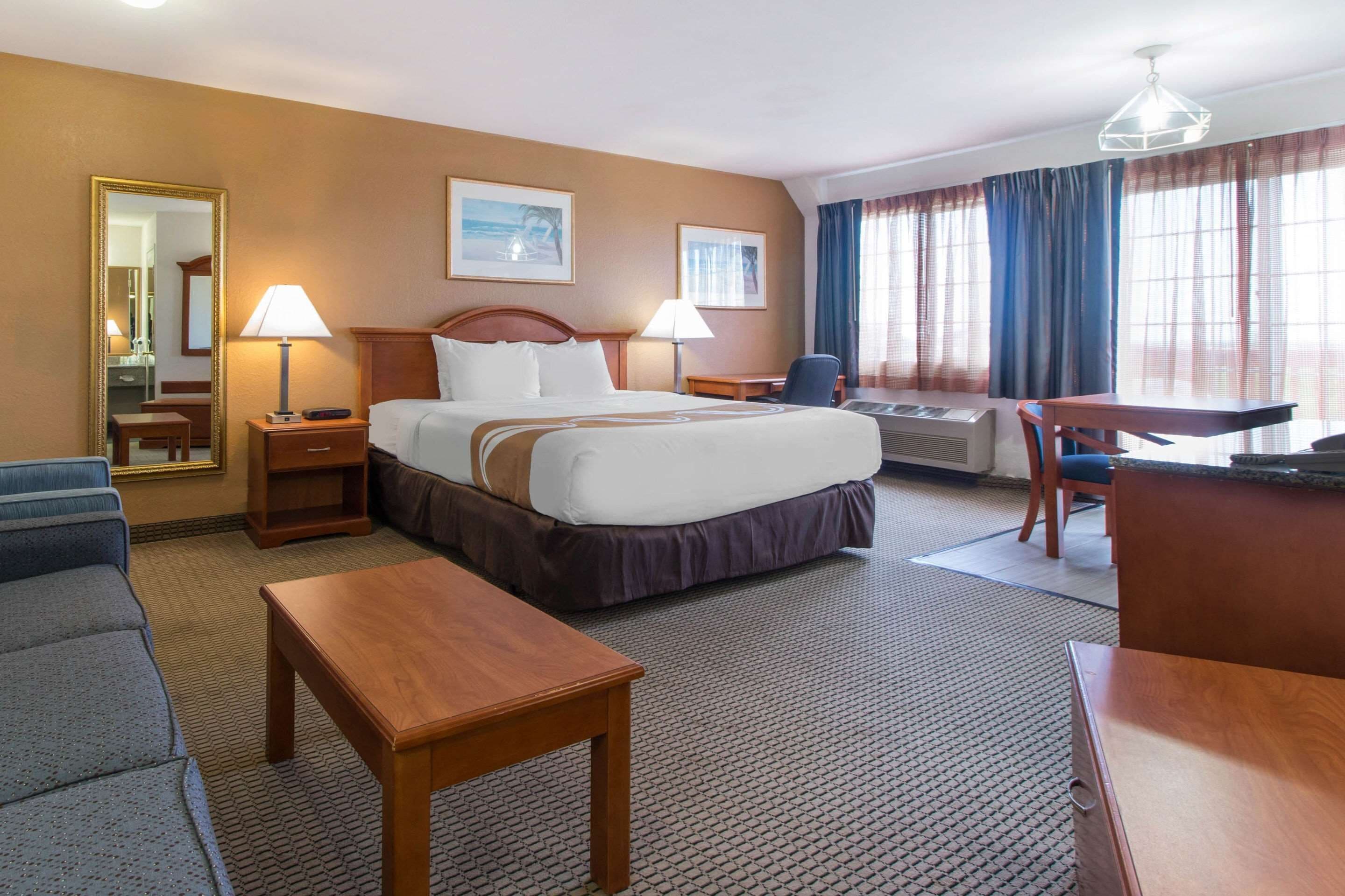 A hotel room with a bed, two bedside tables, a desk, a chair, a sofa, a lamp, a mirror, and a window with curtains.