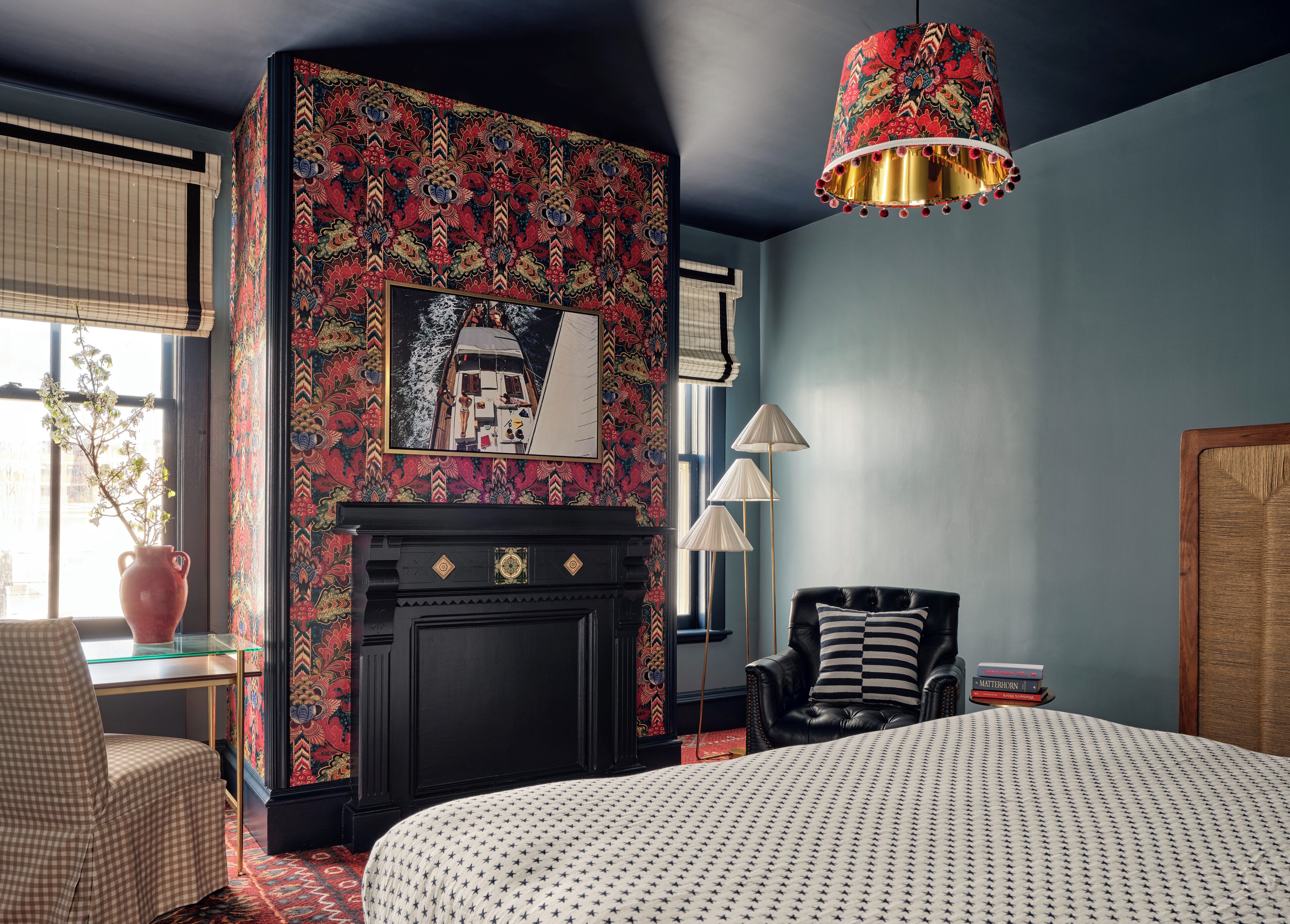 Una habitación con papel tapiz estampado, una chimenea, una cama con colcha estampada, una silla, una mesa con un jarrón y una lámpara colgante.