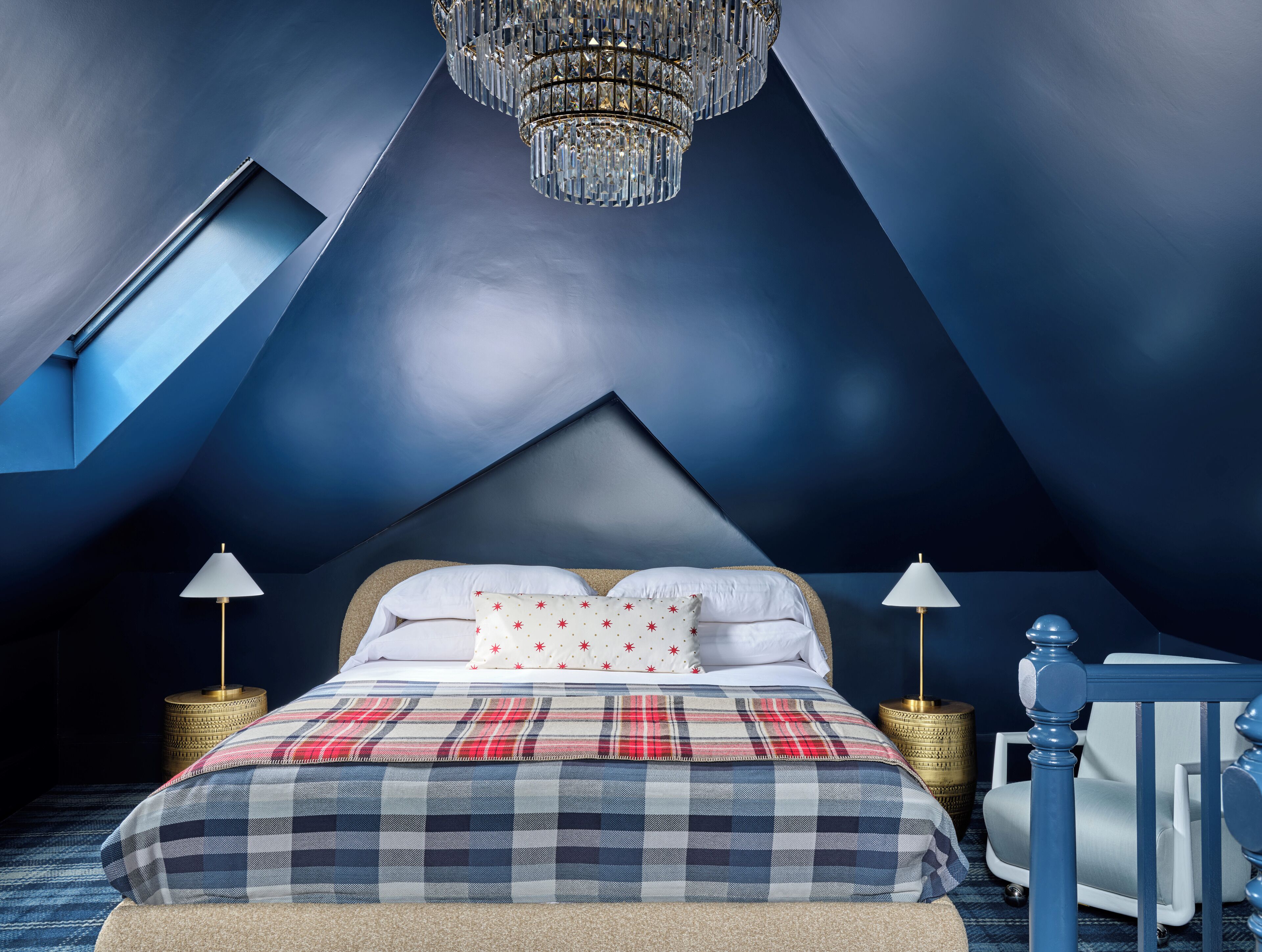 Ein modernes Schlafzimmer mit schrägem Dach, einem großen Bett mit Karo-Bettwäsche, zwei Nachttischlampen und einem Kronleuchter.