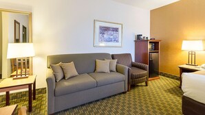 A hotel room with a sofa, armchair, a small table with a lamp, a desk with a lamp, a refrigerator, and a bed.