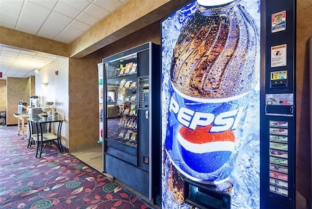 A vending machine with a Pepsi logo and a snack vending machine in a hotel lobby with a patterned carpet and a table with chairs.
