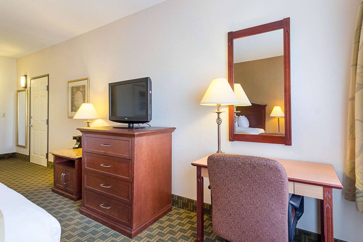 Habitación de hotel con cama, cómoda de madera, televisión, dos lámparas, escritorio y espejo.