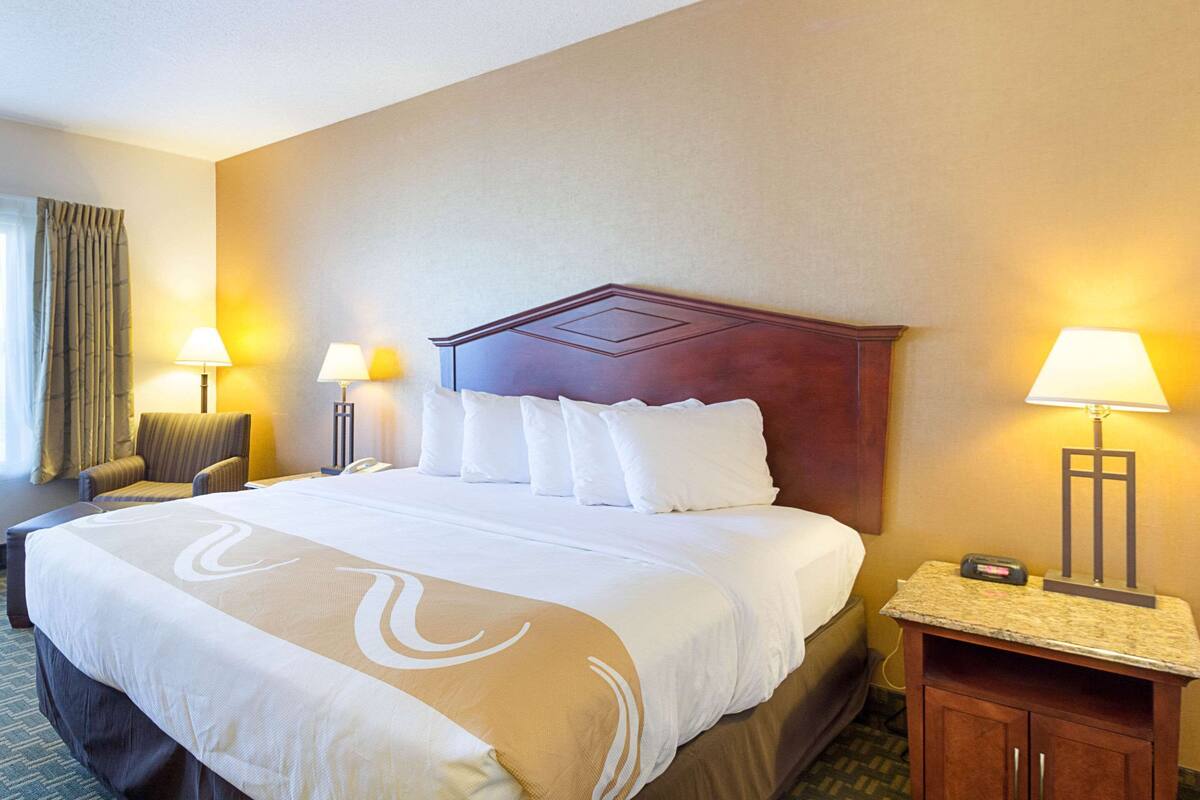 Habitación de hotel con una cama grande, dos lámparas de noche, una silla, una mesita de noche y una ventana con cortinas.