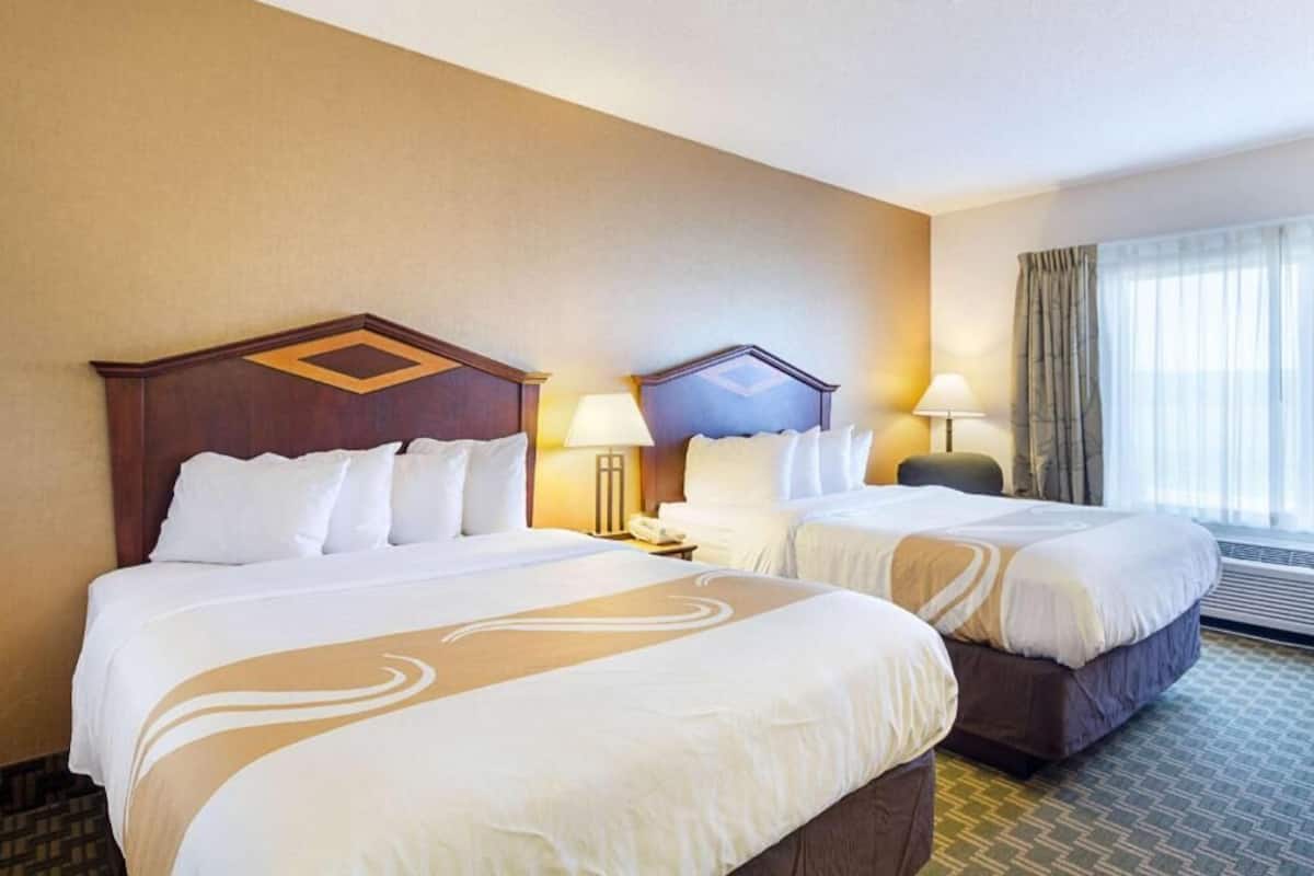 Habitación de hotel con dos camas, una mesita de noche con lámpara, una ventana con cortinas y un aire acondicionado instalado en la pared.