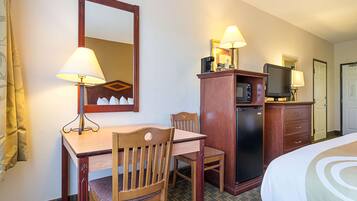 A hotel room with a wooden desk, two chairs, a bed, a television, a coffee maker, and a microwave.