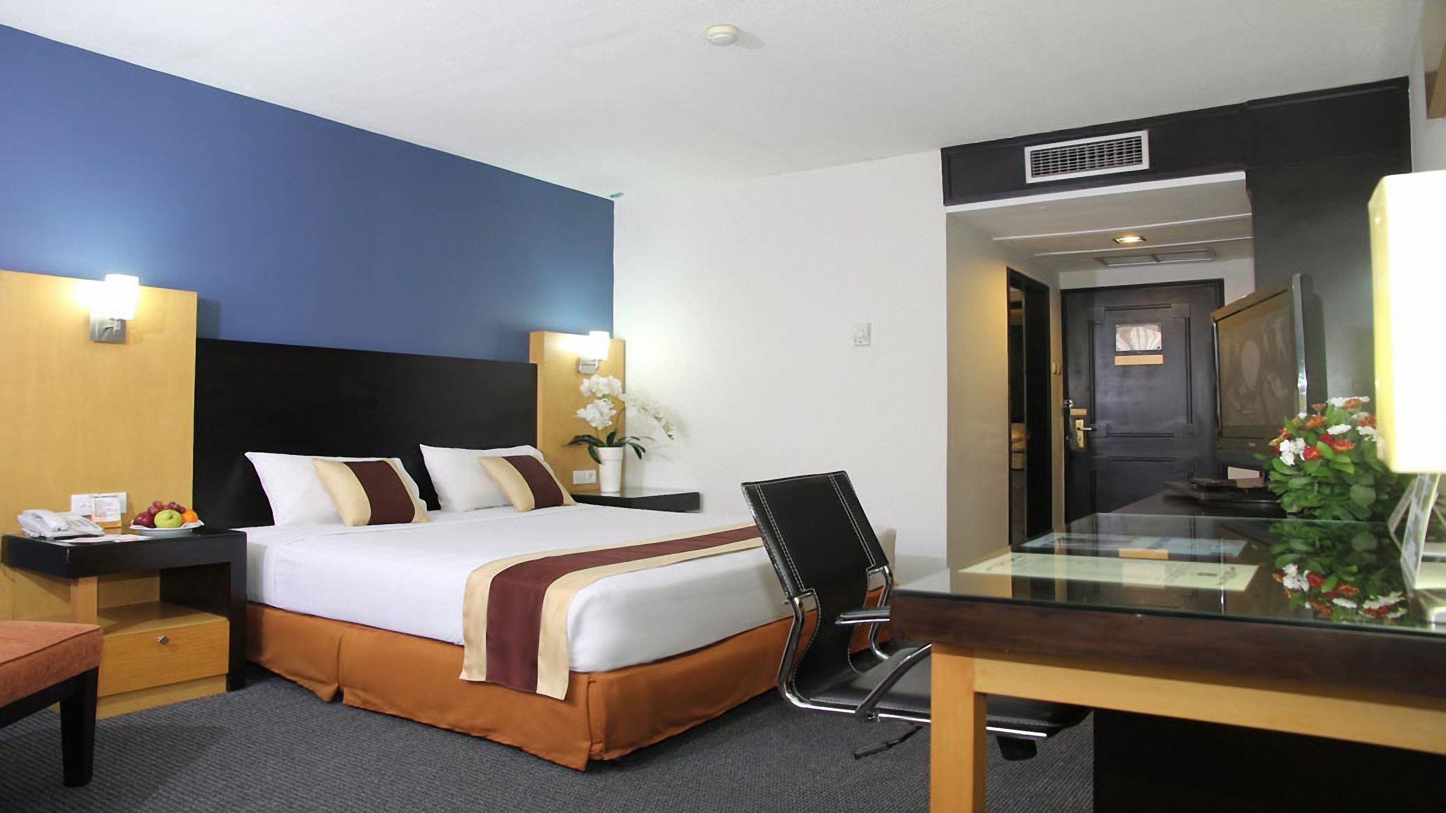 A hotel room with a bed, bedside tables, a desk, a chair, a mirror, and a potted plant.