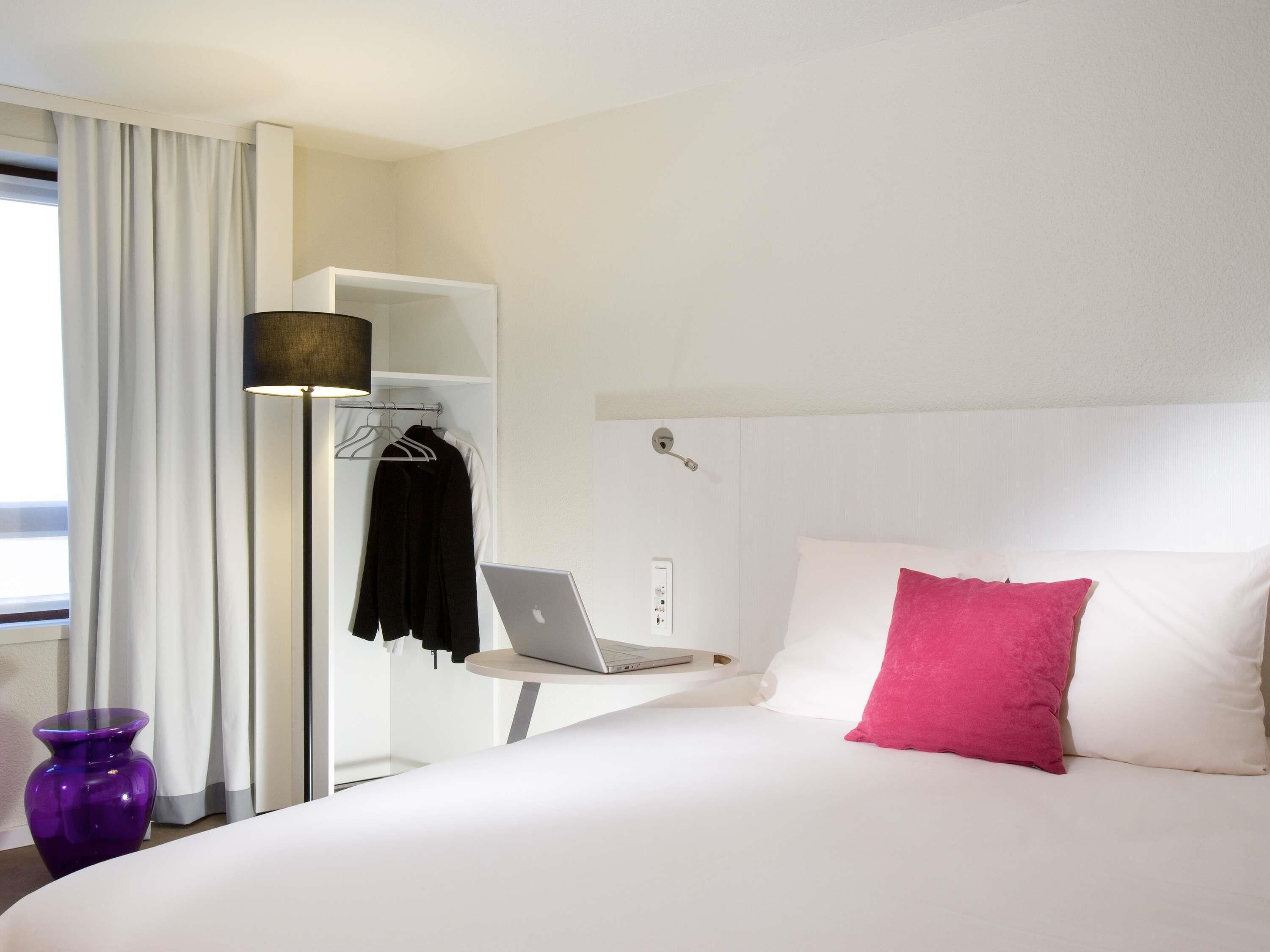 A hotel room with a bed, a laptop on a small table, a wardrobe, and a window with curtains.