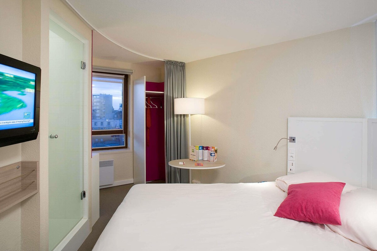 A hotel room with a bed, a TV mounted on the wall, a bedside table with a lamp, and a view of the city through the window.