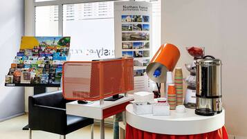 A modern room with a desk, a chair, a table with a red skirt, a coffee machine, and a display of brochures.