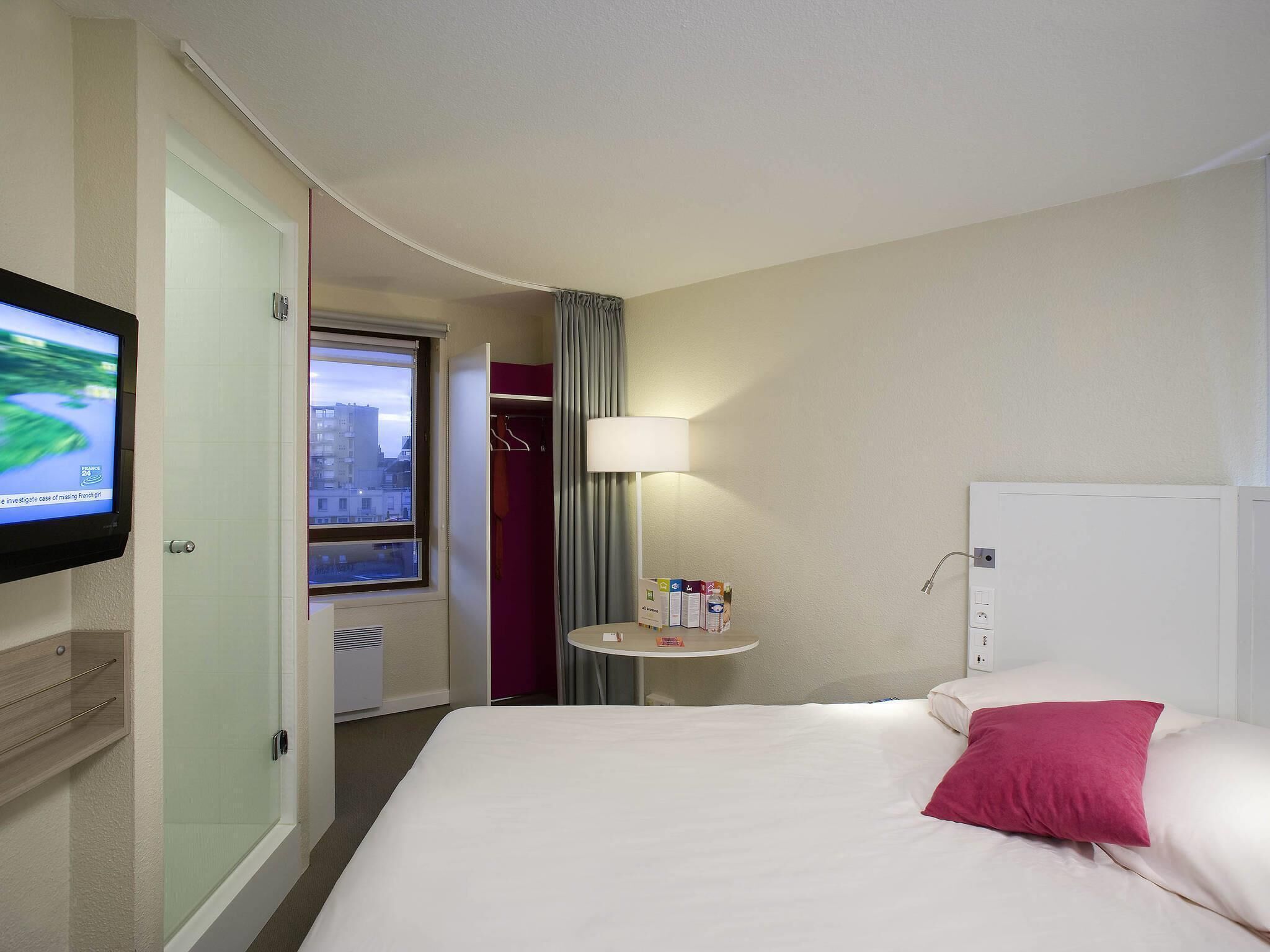 A hotel room with a bed, a TV, a bedside table with a lamp, a closet, and a window with a view of buildings.