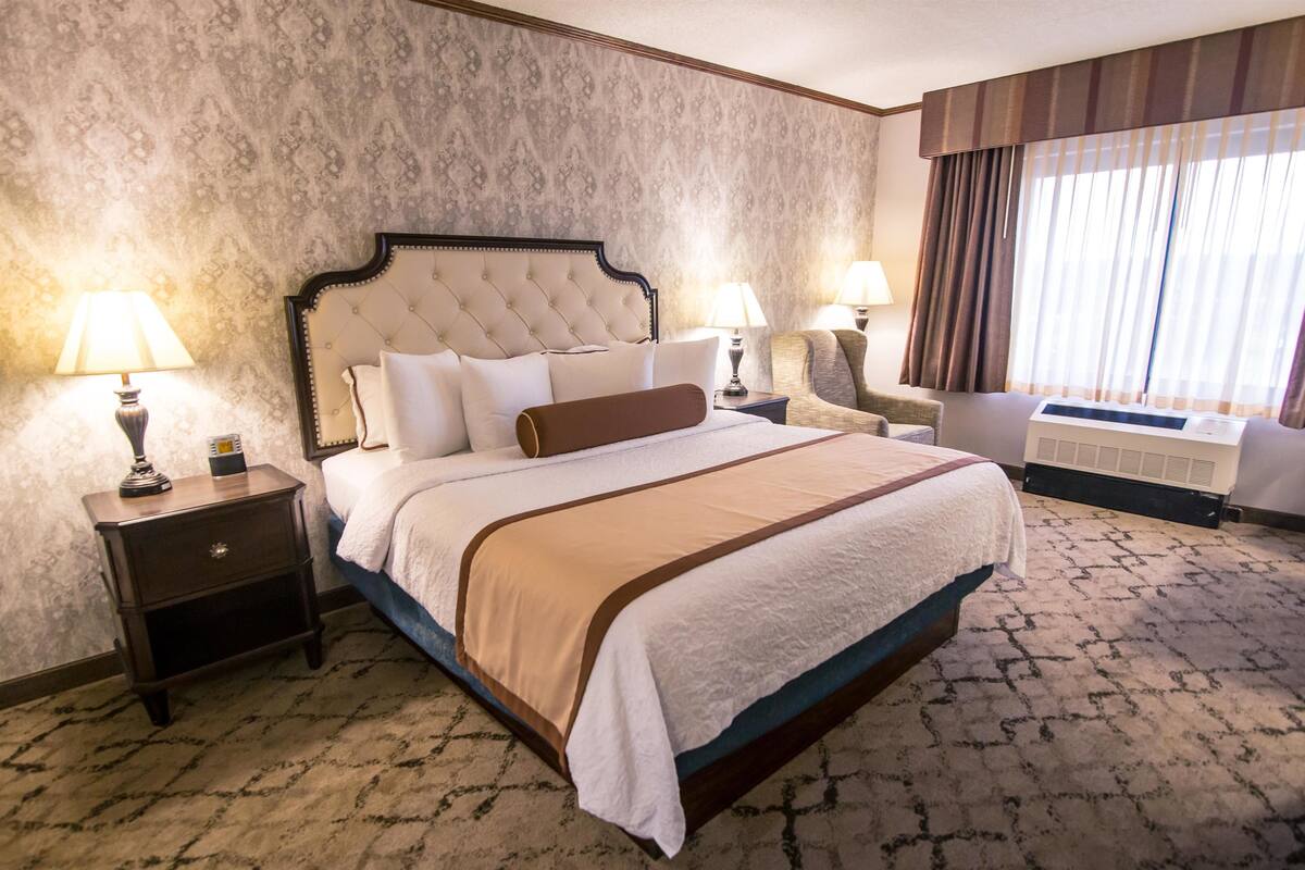 Habitación de hotel con una cama grande, dos mesitas de noche con lámparas, una silla y un amplio ventanal con cortinas.