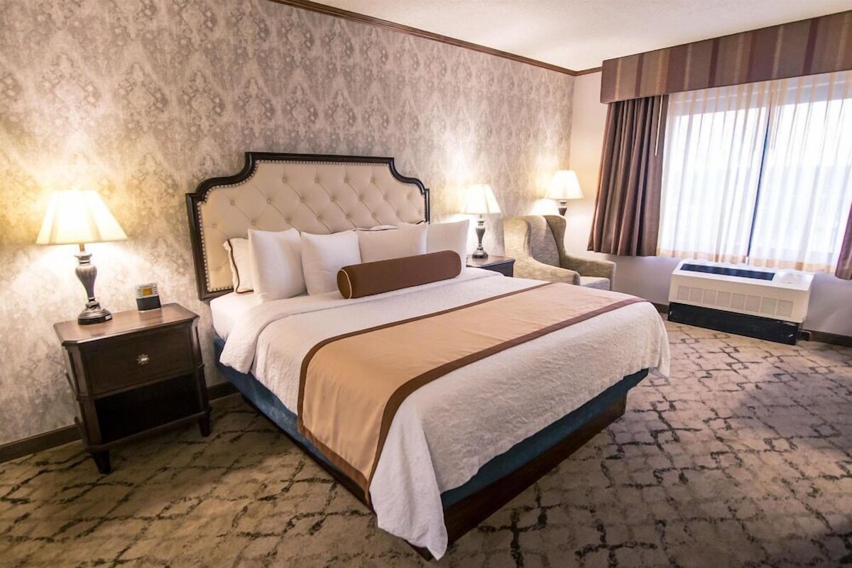 A hotel room with a large bed, two bedside tables with lamps, a chair, and a large window with curtains.