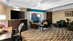 A hotel room with a flat-screen TV, a desk with a lamp, a sofa, a chair, and a patterned carpet.