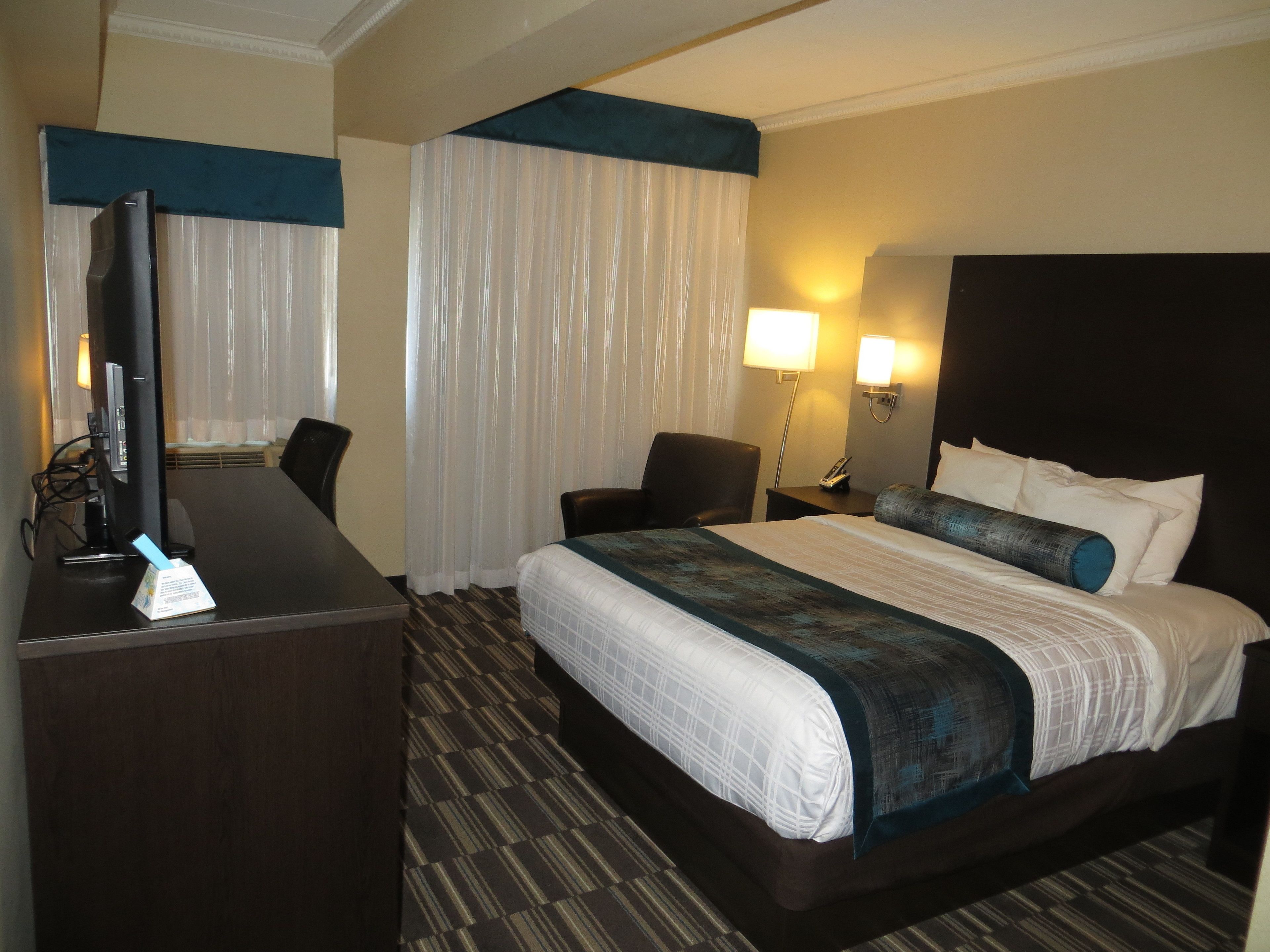 A hotel room with a large bed, a desk with a computer, a chair, a lamp, and a nightstand with a phone.