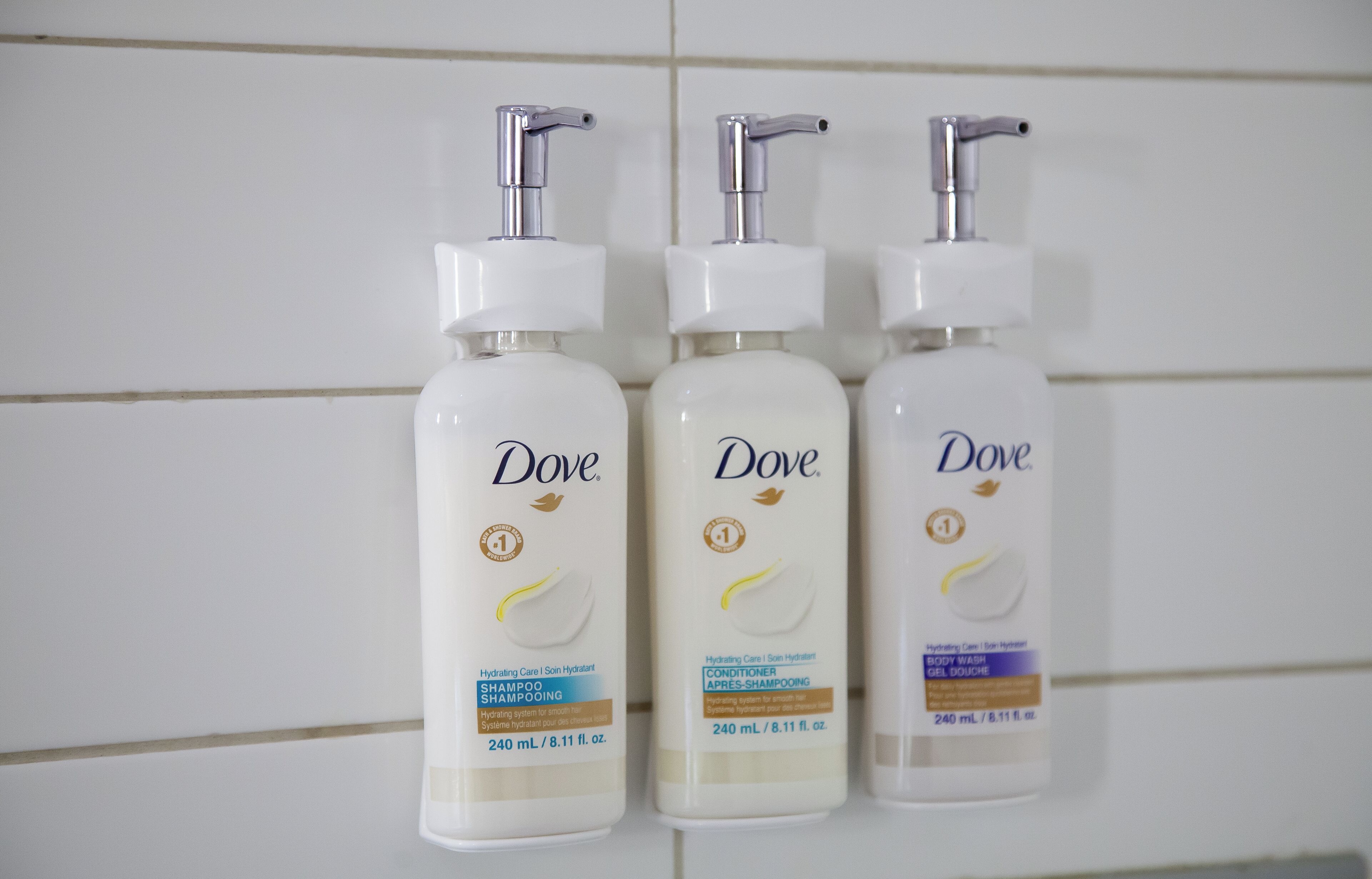 Trois flacons de produits de soins pour la peau de la marque Dove, placés devant un mur carrelé.