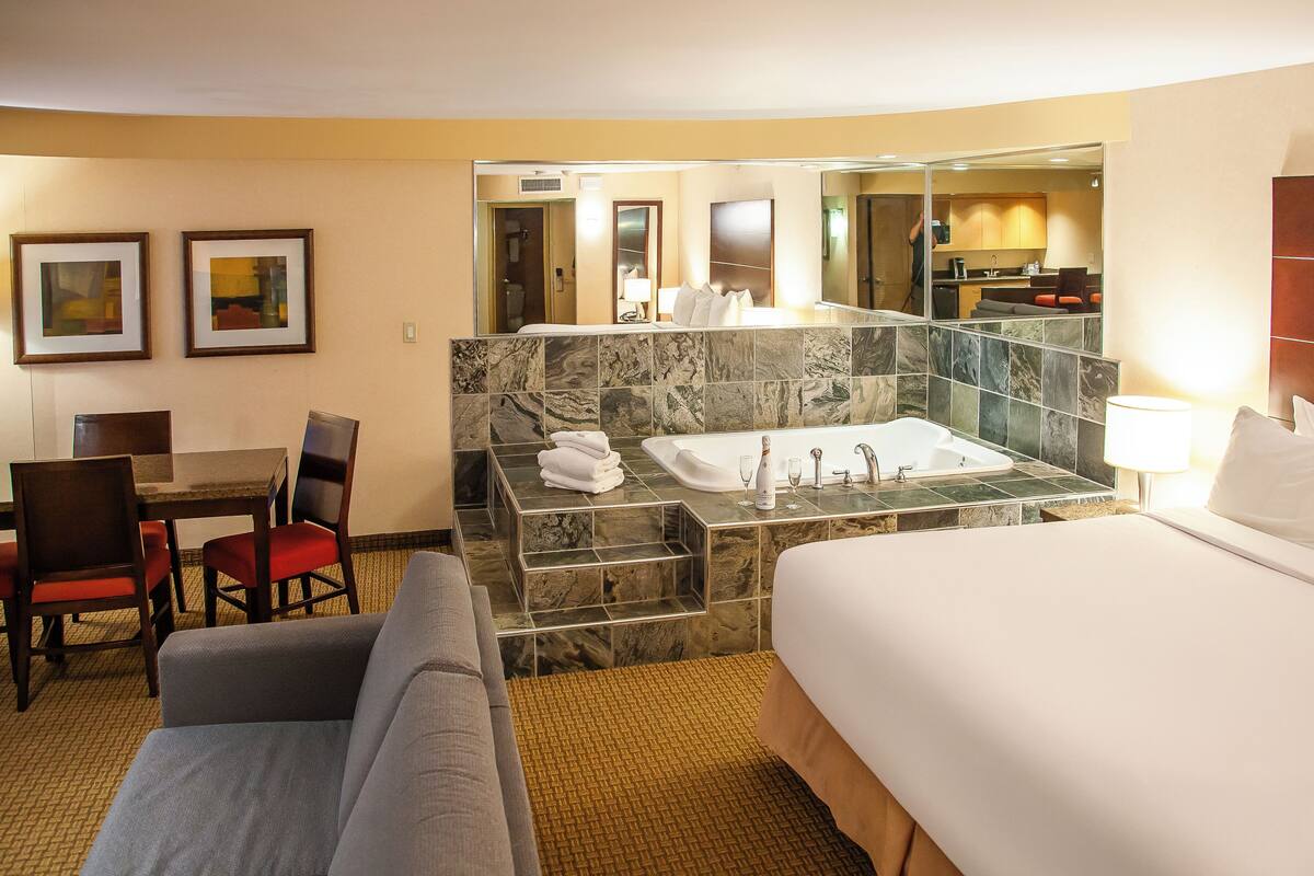 A hotel room with a bed, a sitting area with a table and chairs, a bathroom with a bathtub and a shower, and a dining area with a table and chairs.