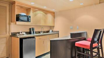 A compact kitchen with a microwave, coffee maker, and a refrigerator. There is a small bar area with a chair.