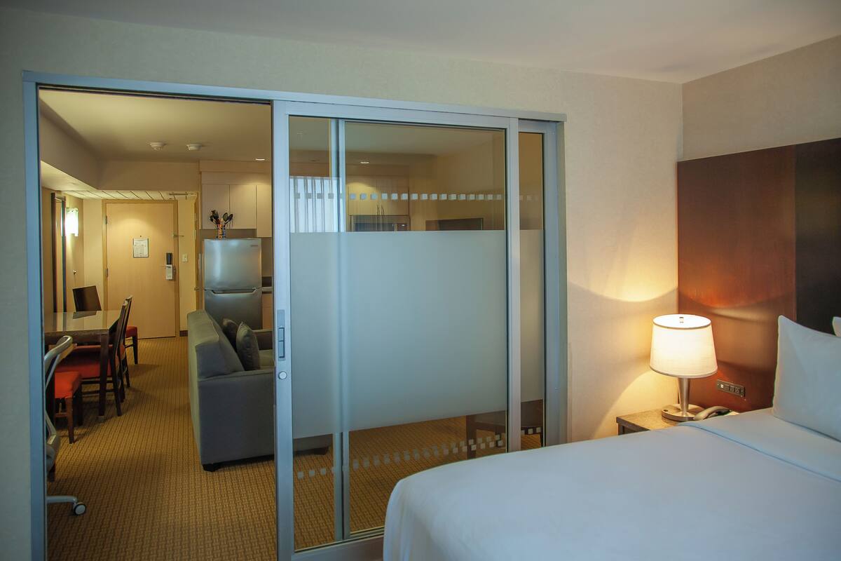 A hotel room with a bed, a desk, a chair, a sofa, and a sliding door leading to a kitchen area.