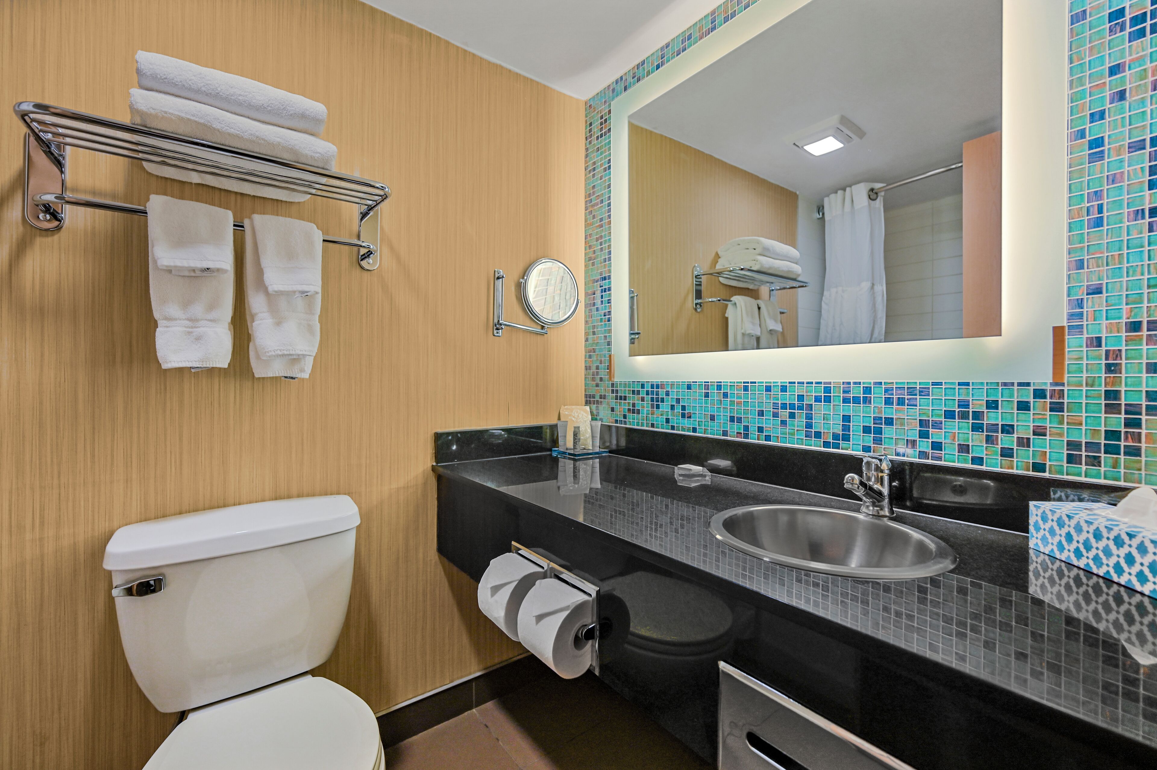 Un baño con inodoro, lavamanos, espejo, toallero y salpicadero de azulejos.