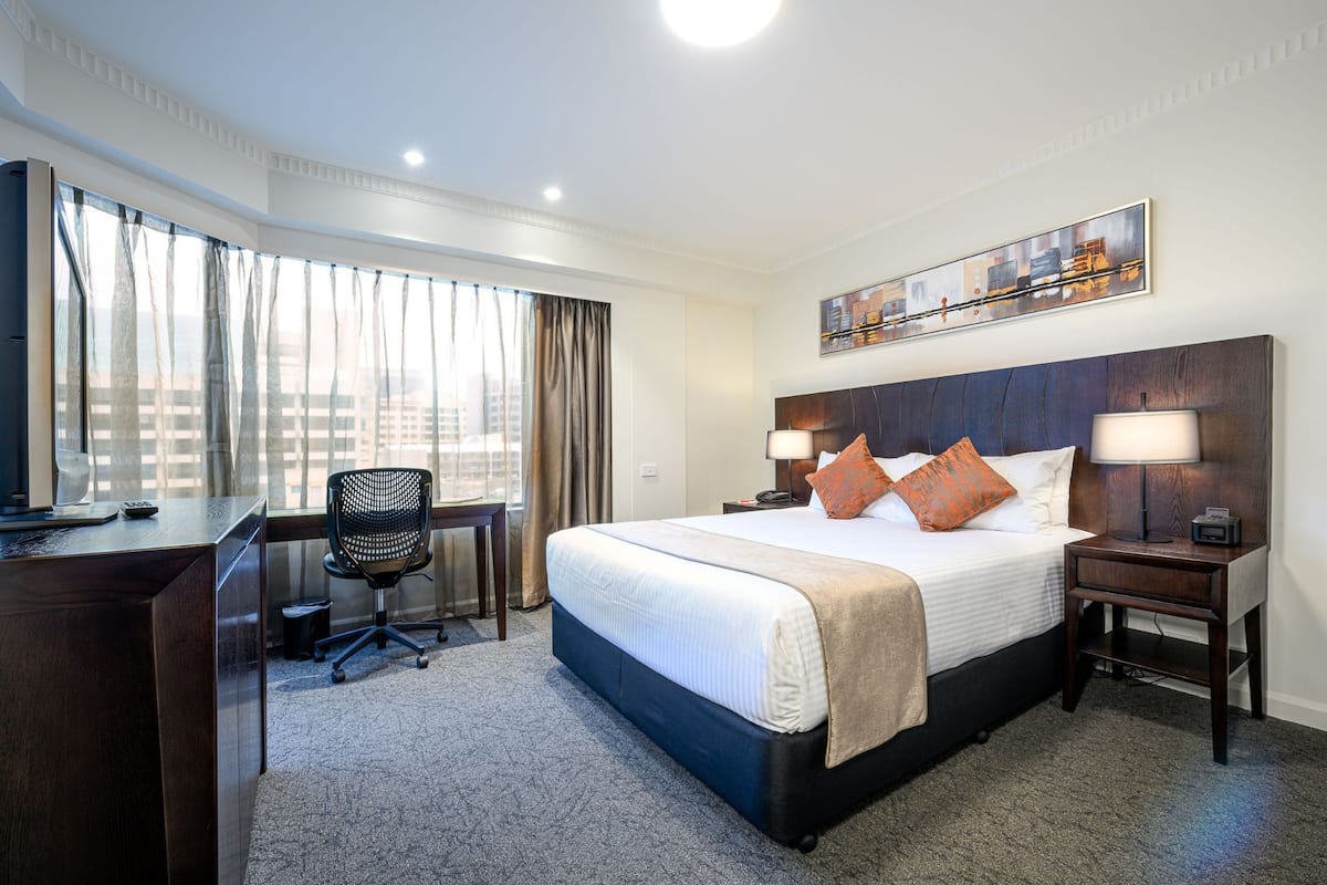 A hotel room with a bed, desk, chair, and a city view through the window.