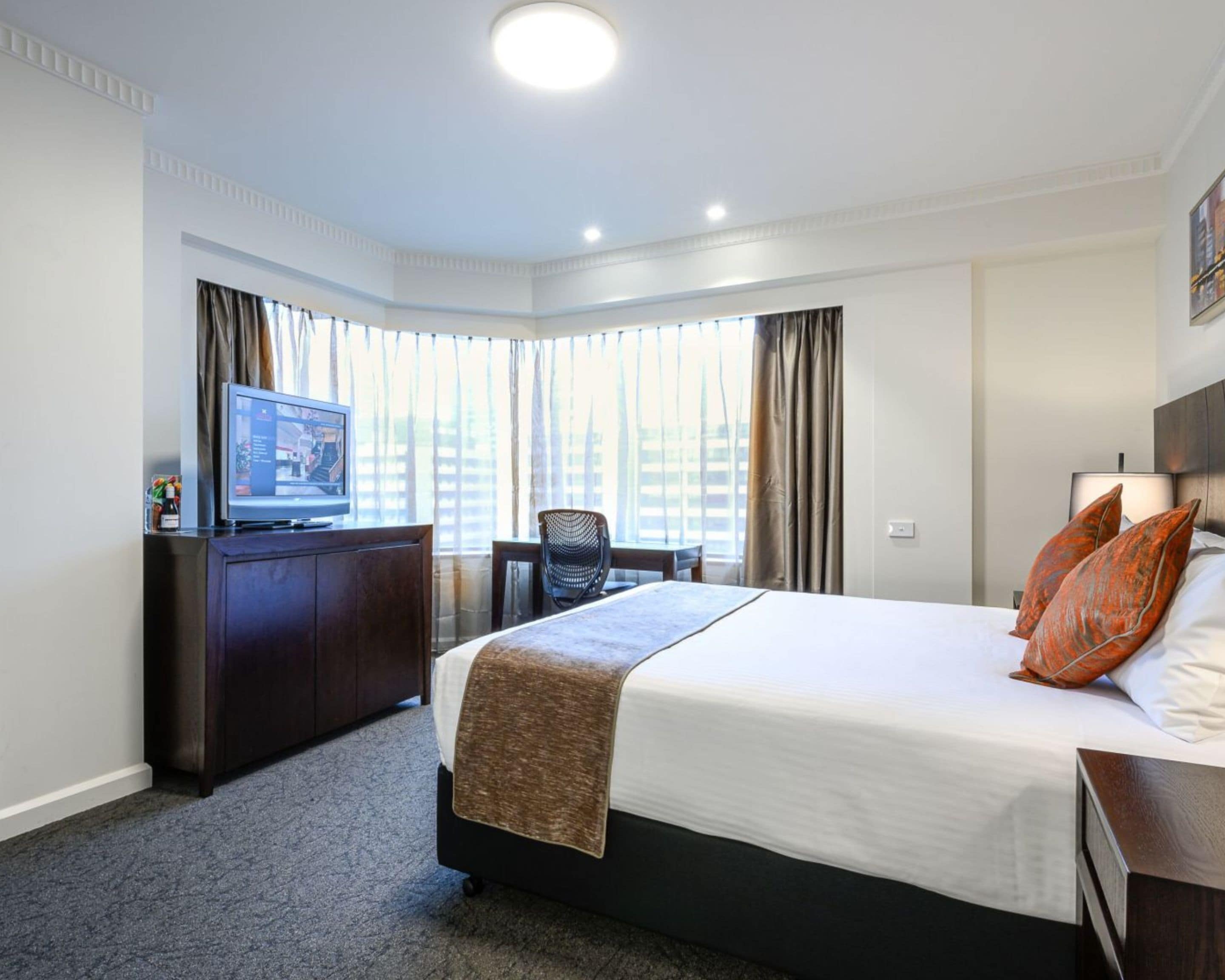 A hotel room with a large bed, a desk with a television, a chair, and a view of buildings through the windows.