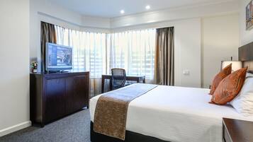 A hotel room with a large bed, a desk with a television, a chair, and a view of buildings through the windows.