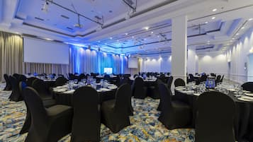 Un salón de conferencias con filas de mesas y sillas, equipado con proyectores y pantallas, y un gran telón de fondo.