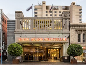 Exterior del Hotel Grand Chancellor, destacando una entrada imponente con fachada de vidrio, dos árboles simétricos al frente y una bandera en la parte superior.