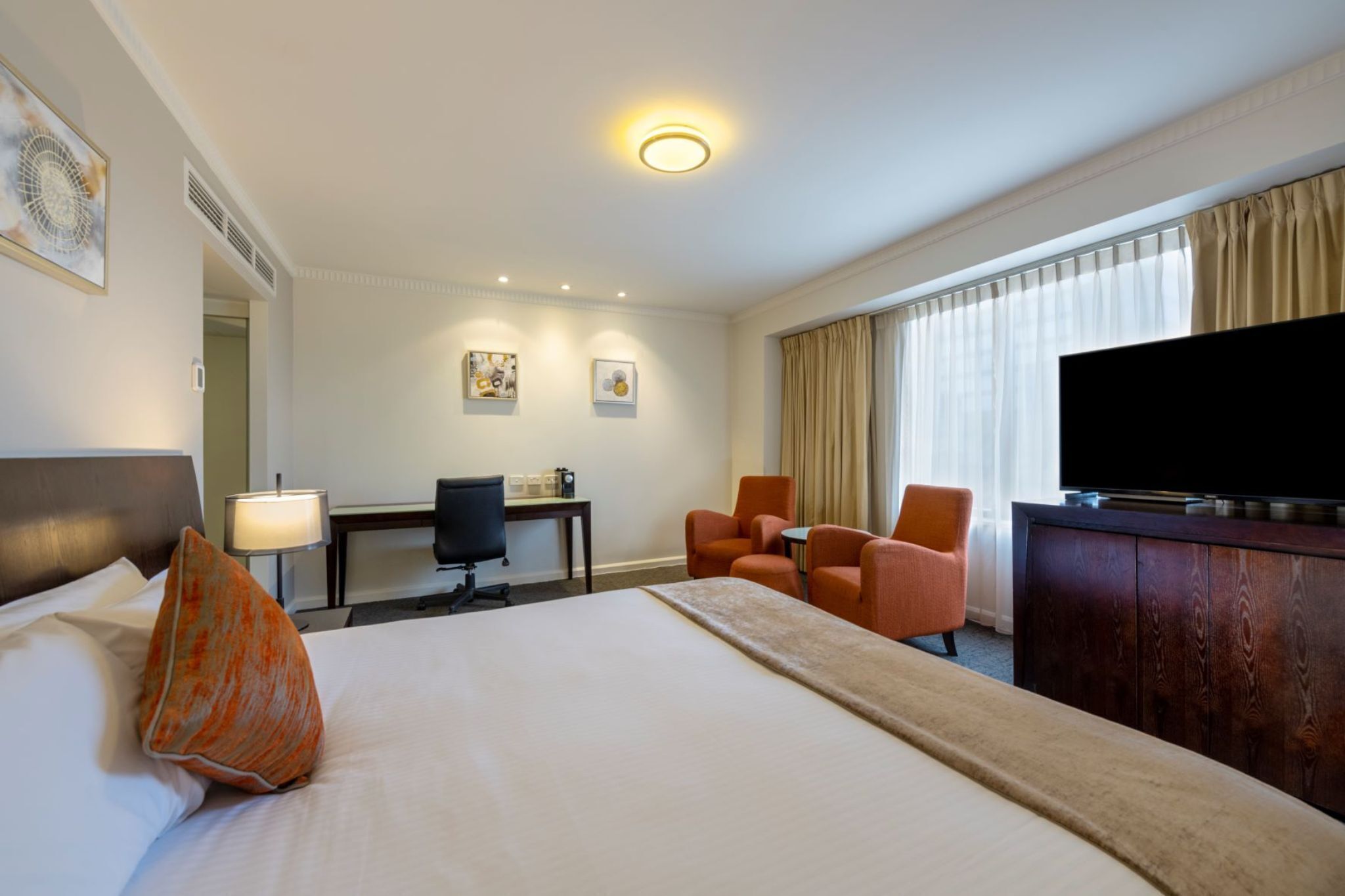 A hotel room with a bed, a desk, two chairs, a television, and a large window with curtains.