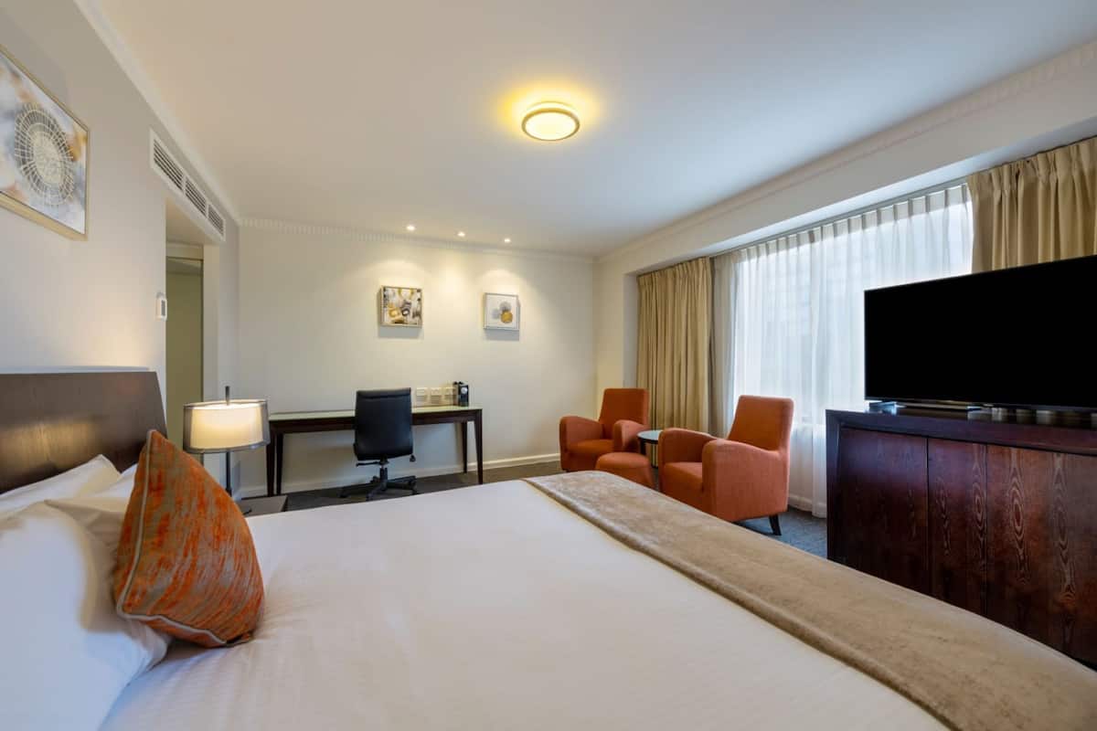 A hotel room with a bed, a desk, two chairs, a television, and a large window with curtains.