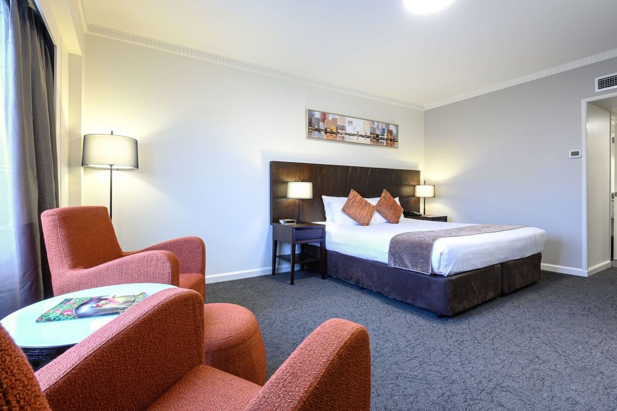 A hotel room with a bed, two bedside tables with lamps, a chair, a small table, and a painting on the wall.