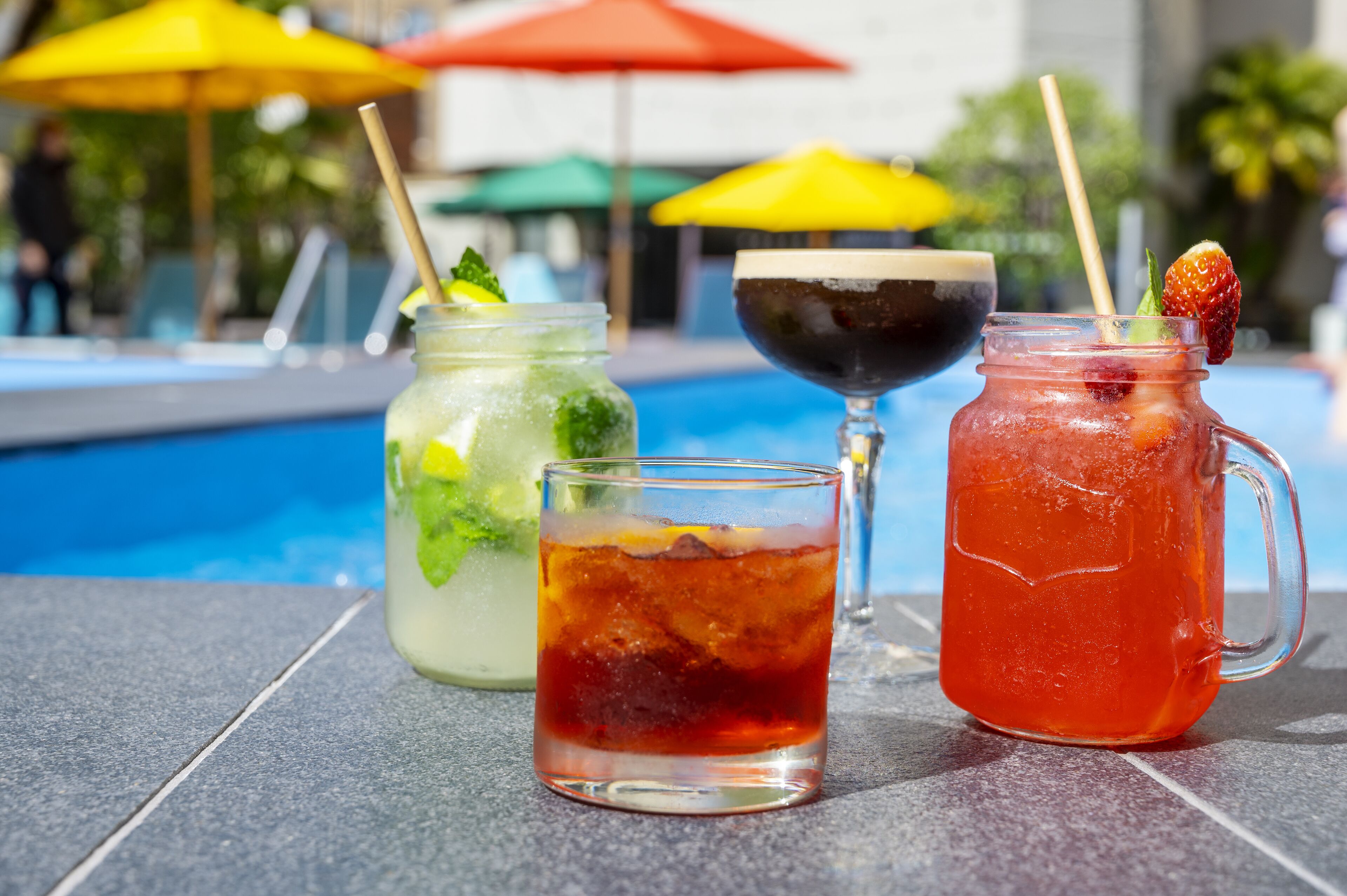 A poolside setting with three drinks: a green cocktail, a red cocktail, and a dark drink in a glass.