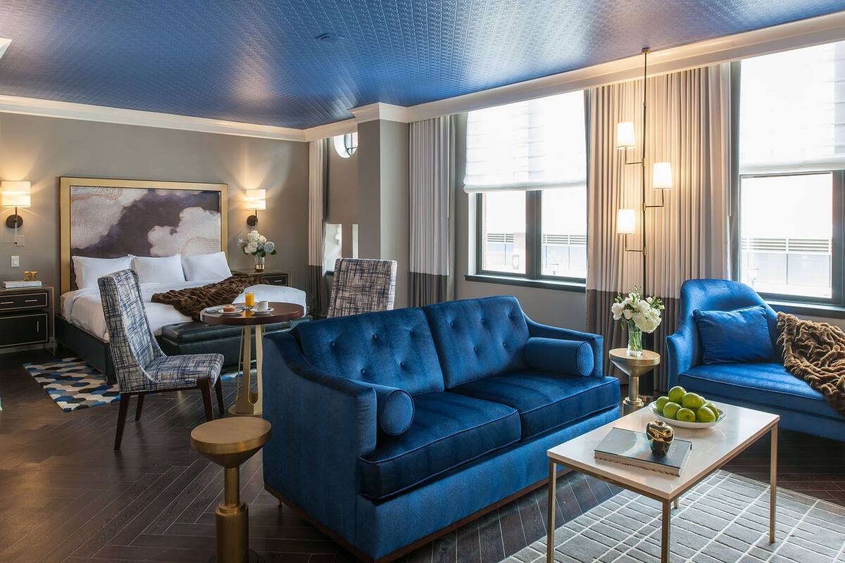 A modern hotel room with a large bed, a blue sofa, a coffee table, and a dining area.