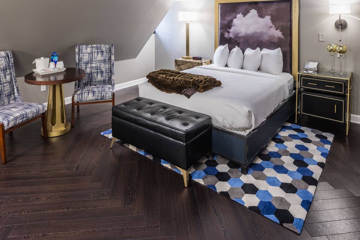 A modern hotel room with a large bed, a small round table, two chairs, a nightstand, and a ceiling with a geometric pattern.