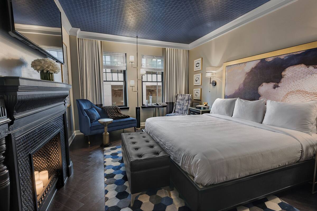 A modern hotel room with a large bed, a blue armchair, a fireplace, and a geometric-patterned rug.