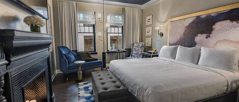A modern hotel room with a large bed, a blue armchair, a fireplace, and a geometric-patterned rug.