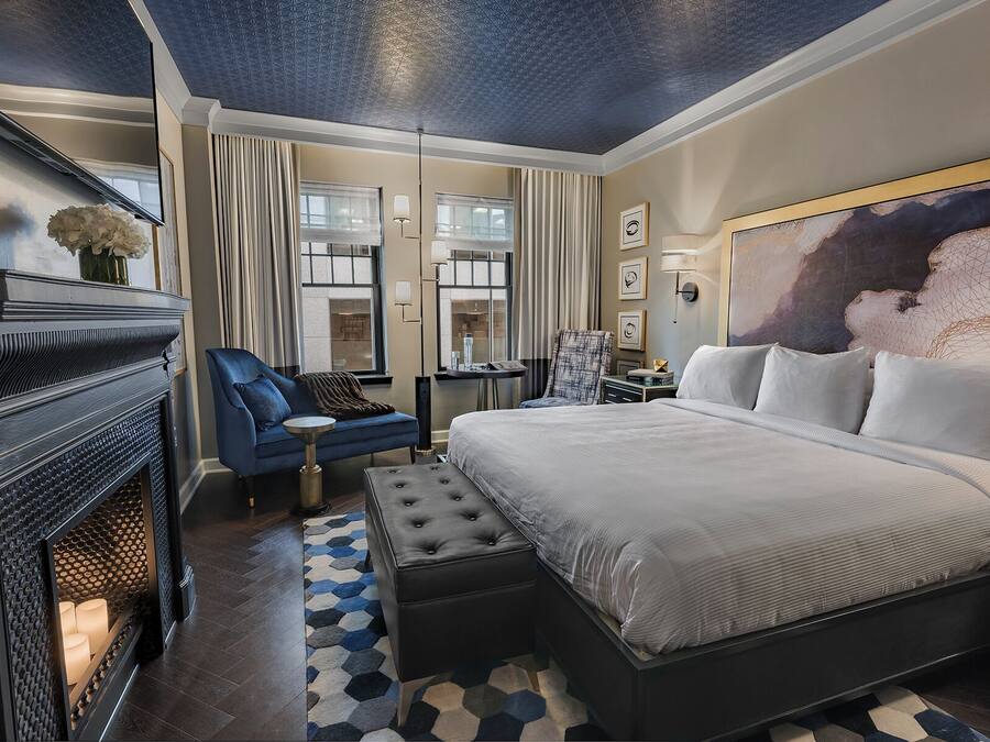 Una habitación de hotel moderna con una cama grande, un sillón azul, una chimenea y una alfombra con motivos geométricos.