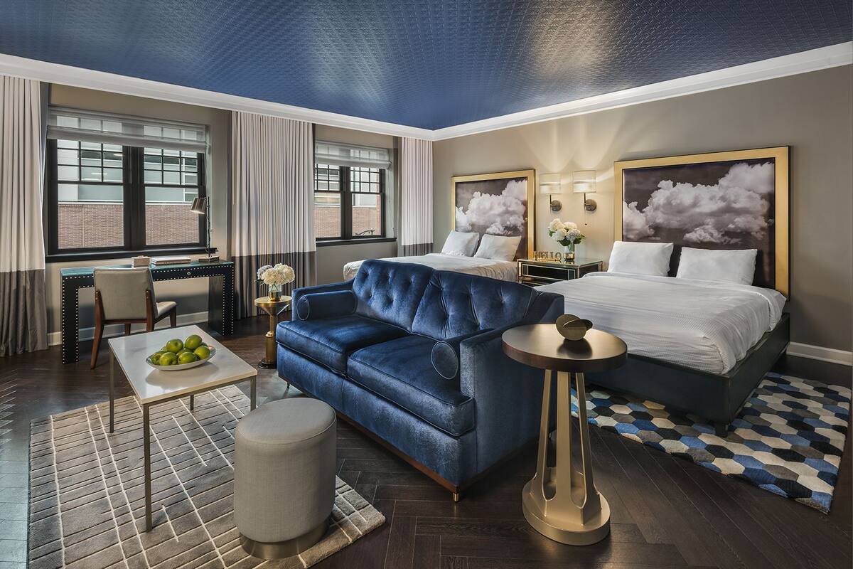 A hotel room with a large bed, a blue sofa, a small table with apples, and a round table with a lamp.