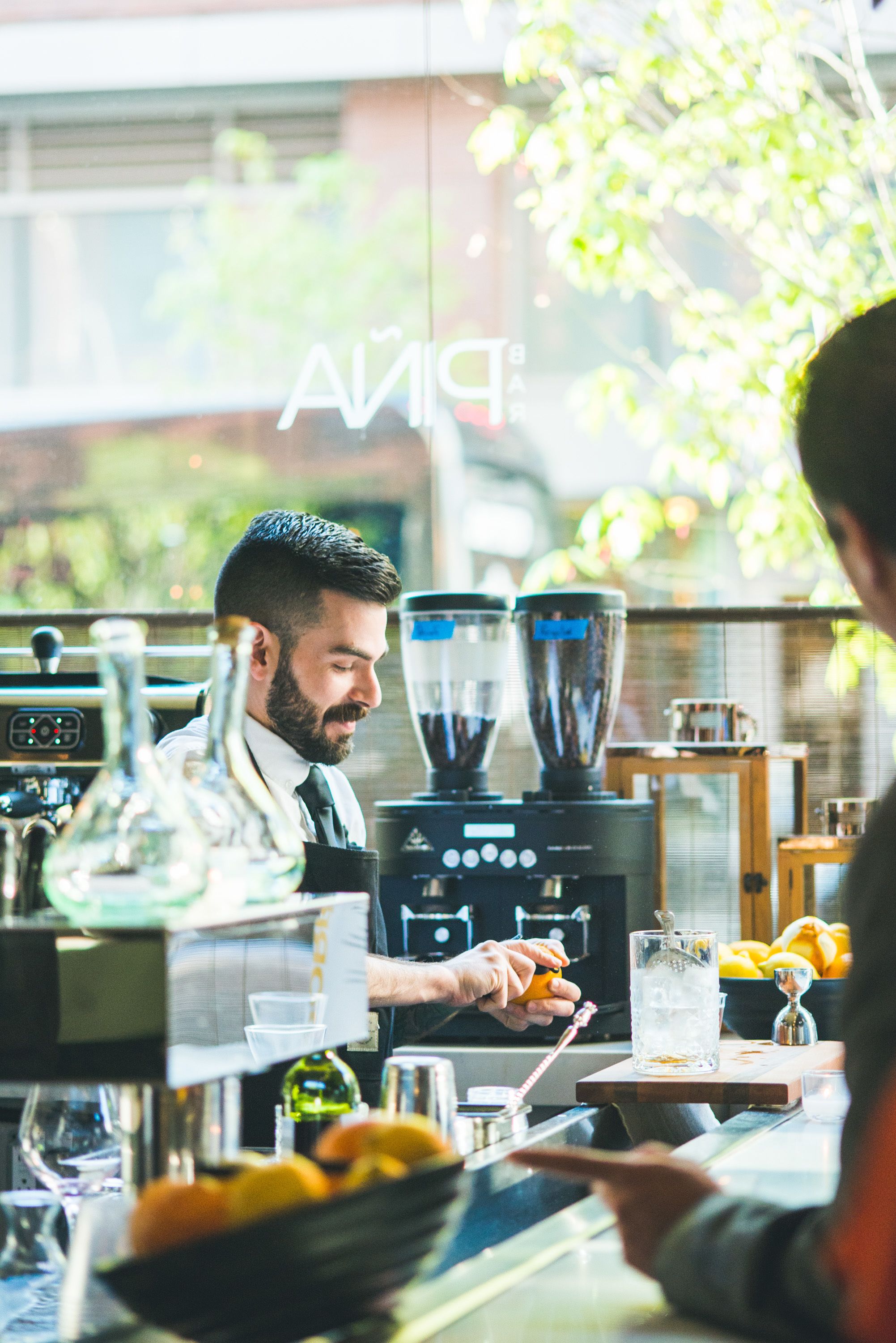 Un bar con un cantinero preparando una bebida, una máquina de café y un vaso de agua.