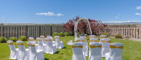 A wedding ceremony setup with white chairs and yellow sashes arranged on a grassy lawn, a wooden fence, and a backdrop of trees and mountains.