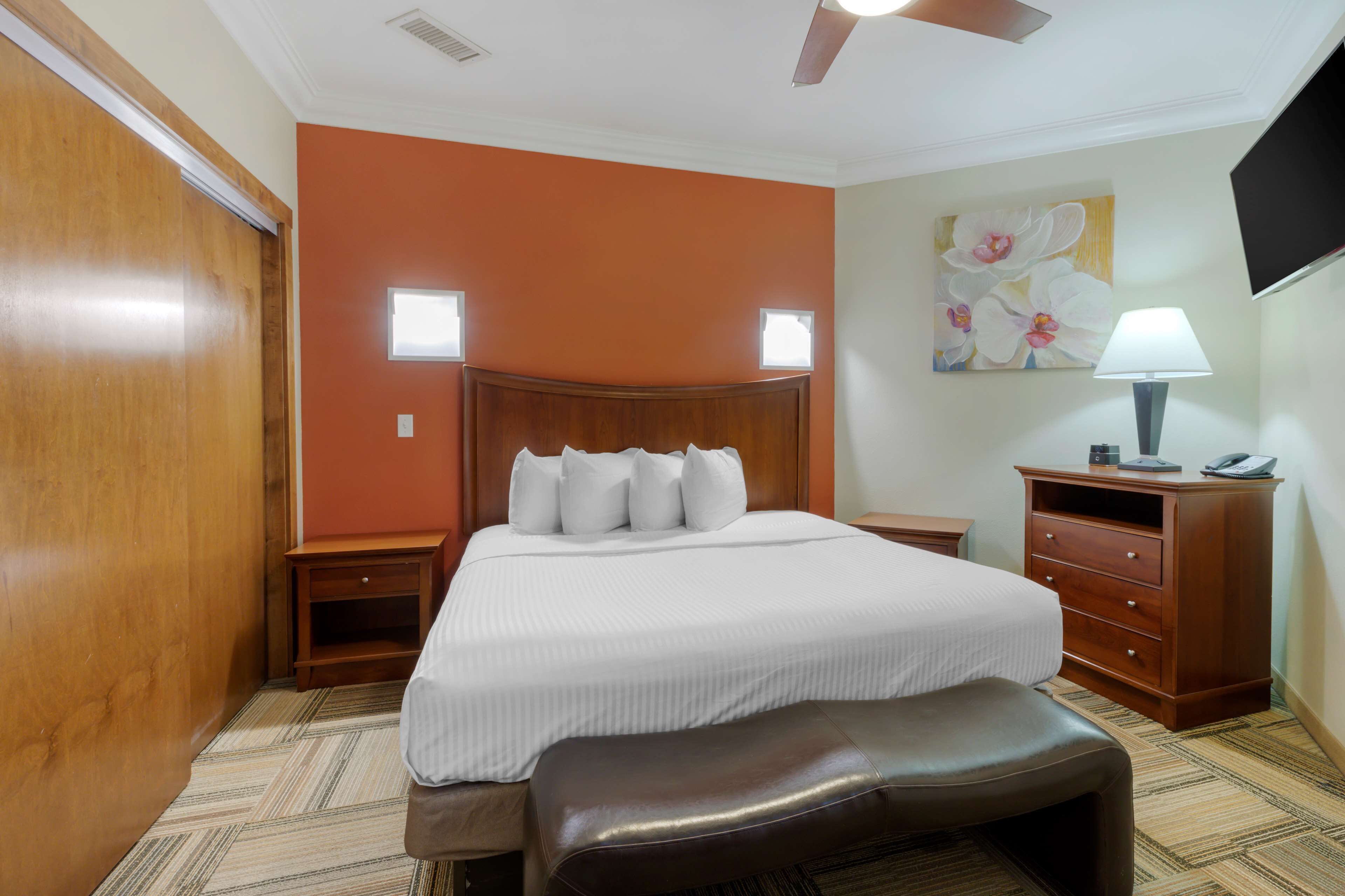 Une chambre d’hôtel avec un grand lit, une tête de lit en bois, un banc, une table de chevet, une commode, une lampe et un tableau au mur.