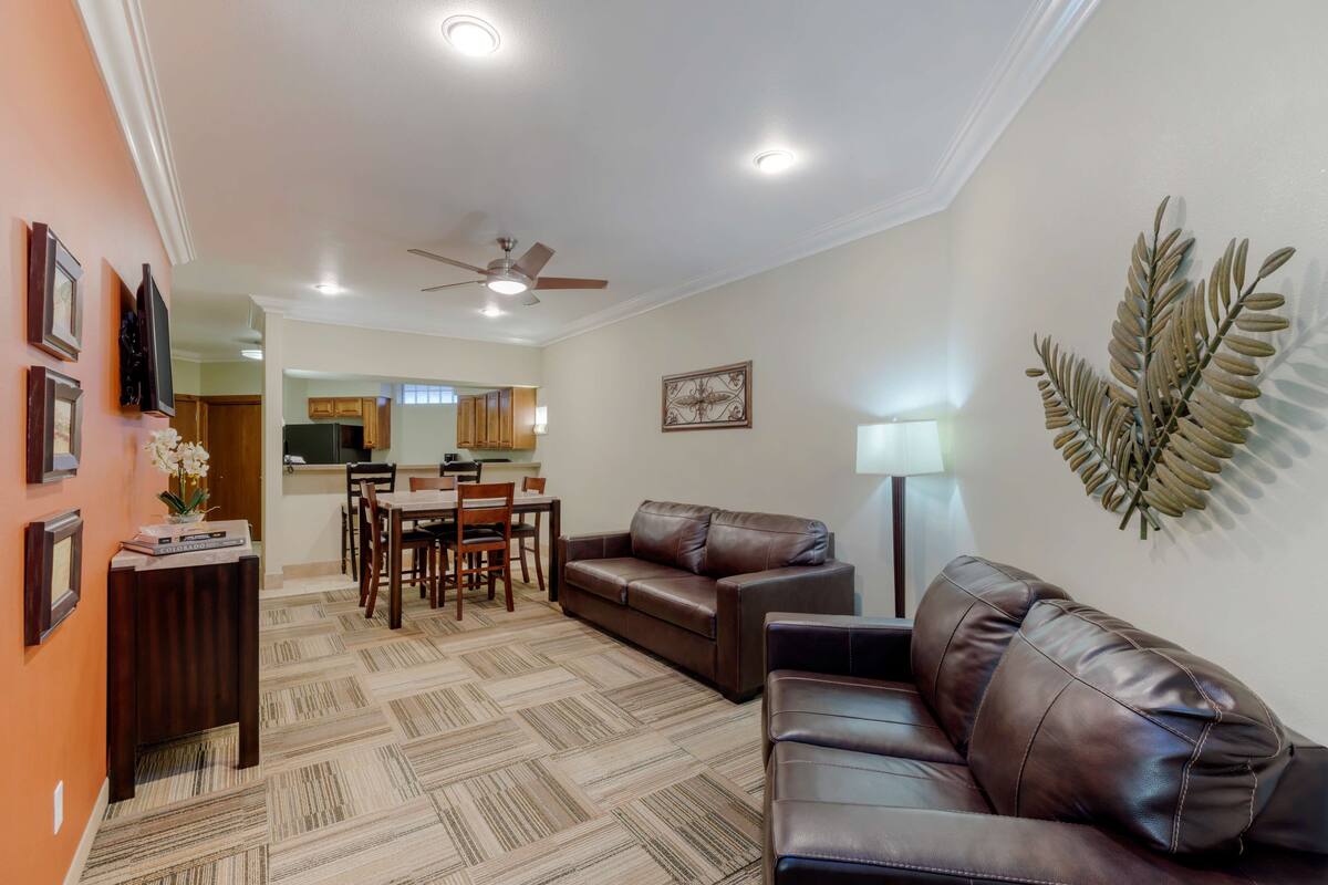 A living room with a brown leather sofa, a dining table with chairs, a ceiling fan, and a decorative leaf wall piece.