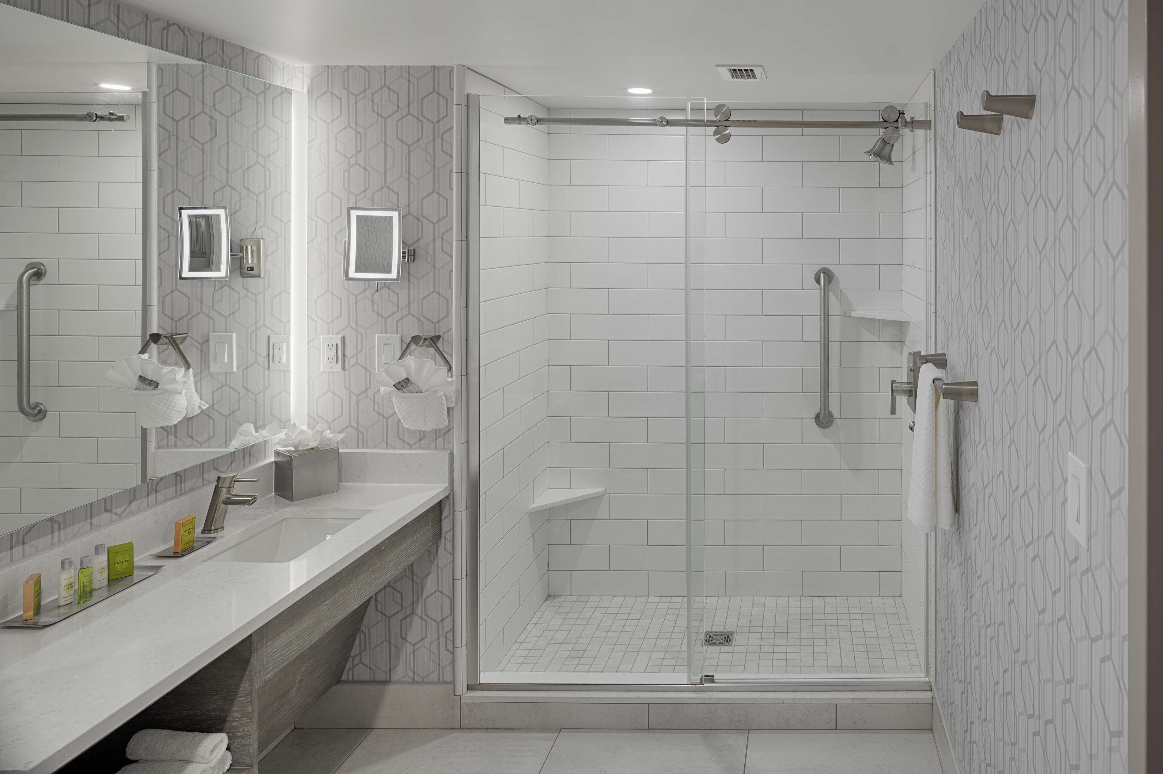 Un baño moderno con un espejo grande, dos lavamanos, zona de ducha y tocador con artículos de aseo.