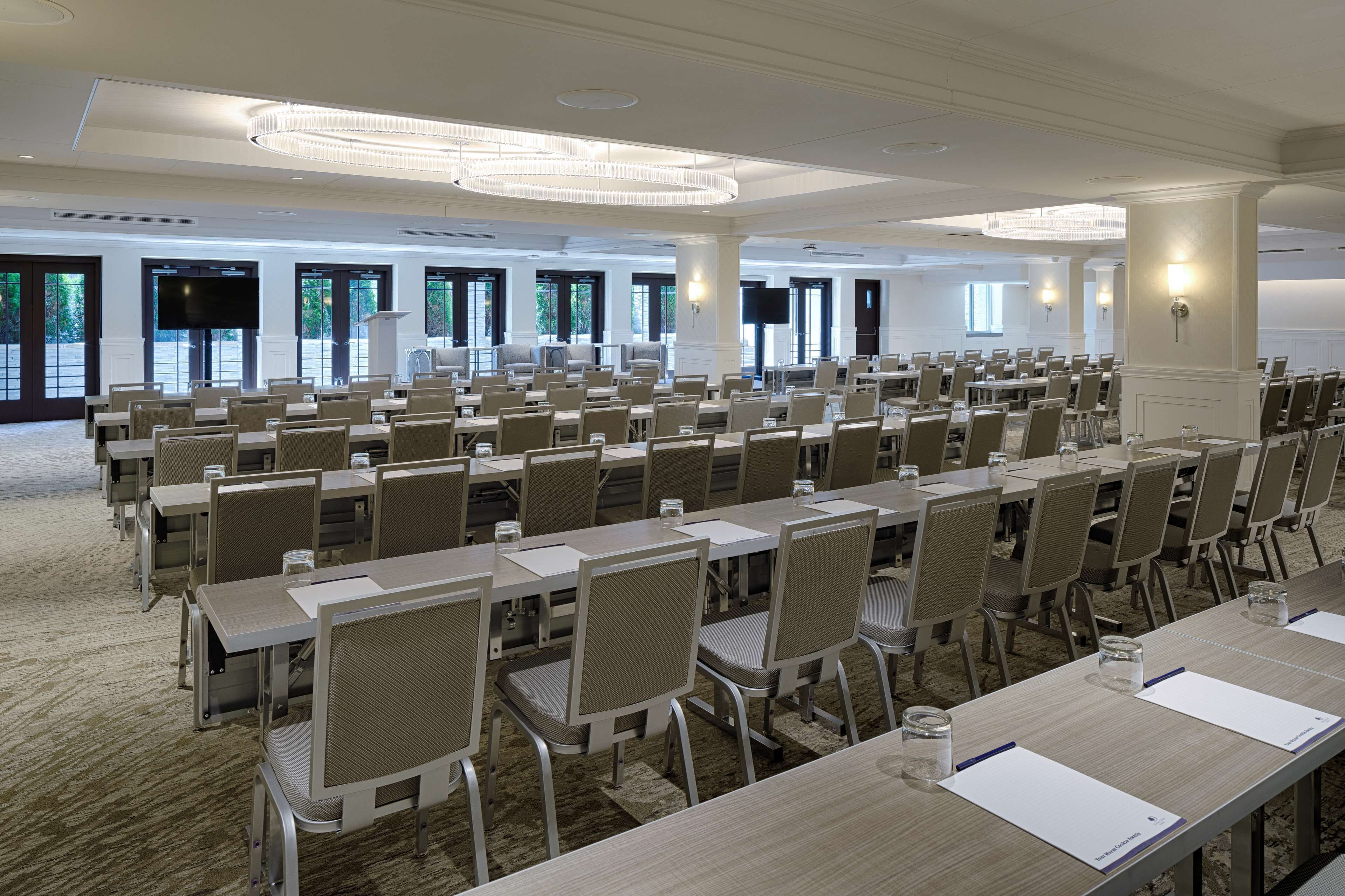 Un salón de conferencias con filas de mesas y sillas, ventanas amplias y una araña.