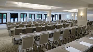 Un salón de conferencias con filas de mesas y sillas, ventanas amplias y una araña.