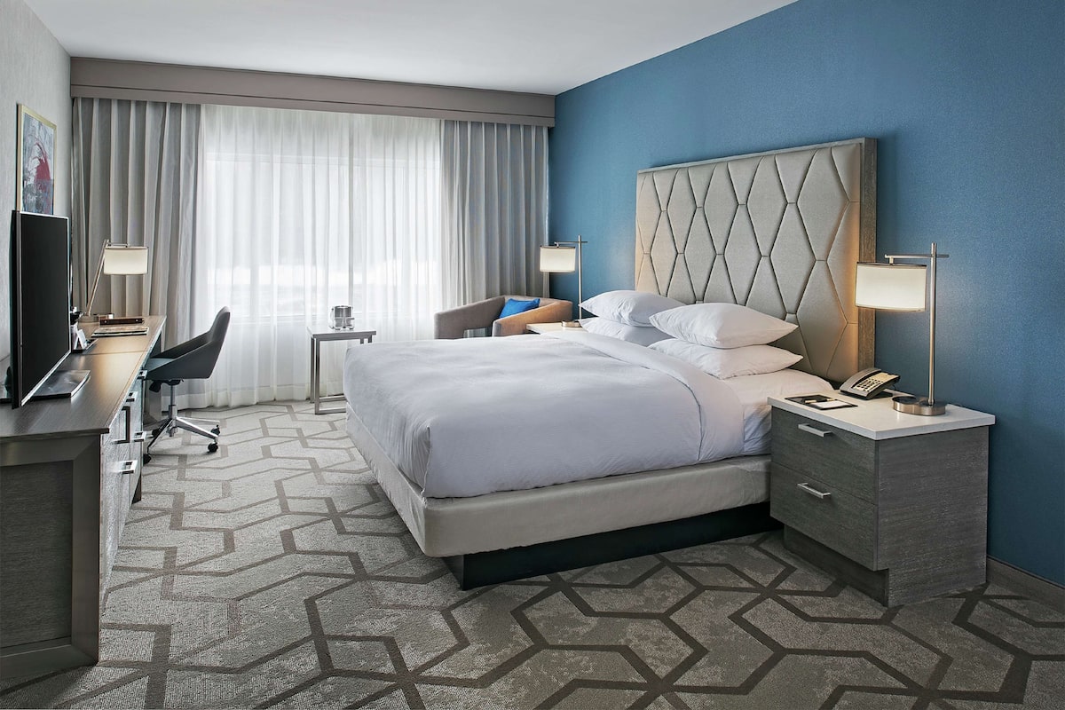 A hotel room with a large bed, a desk with a chair, a TV, and a desk lamp.