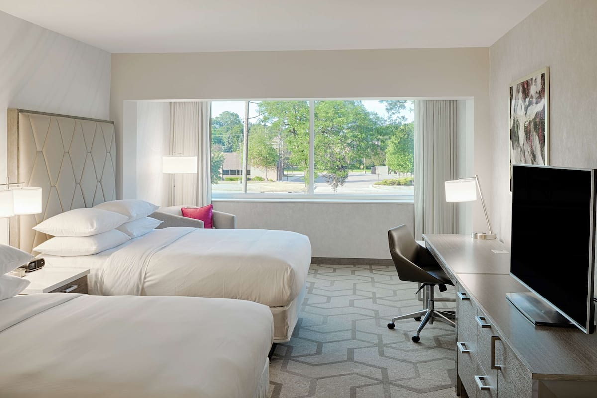 A hotel room with two beds, a desk, a TV, and a window with a view of trees and buildings.