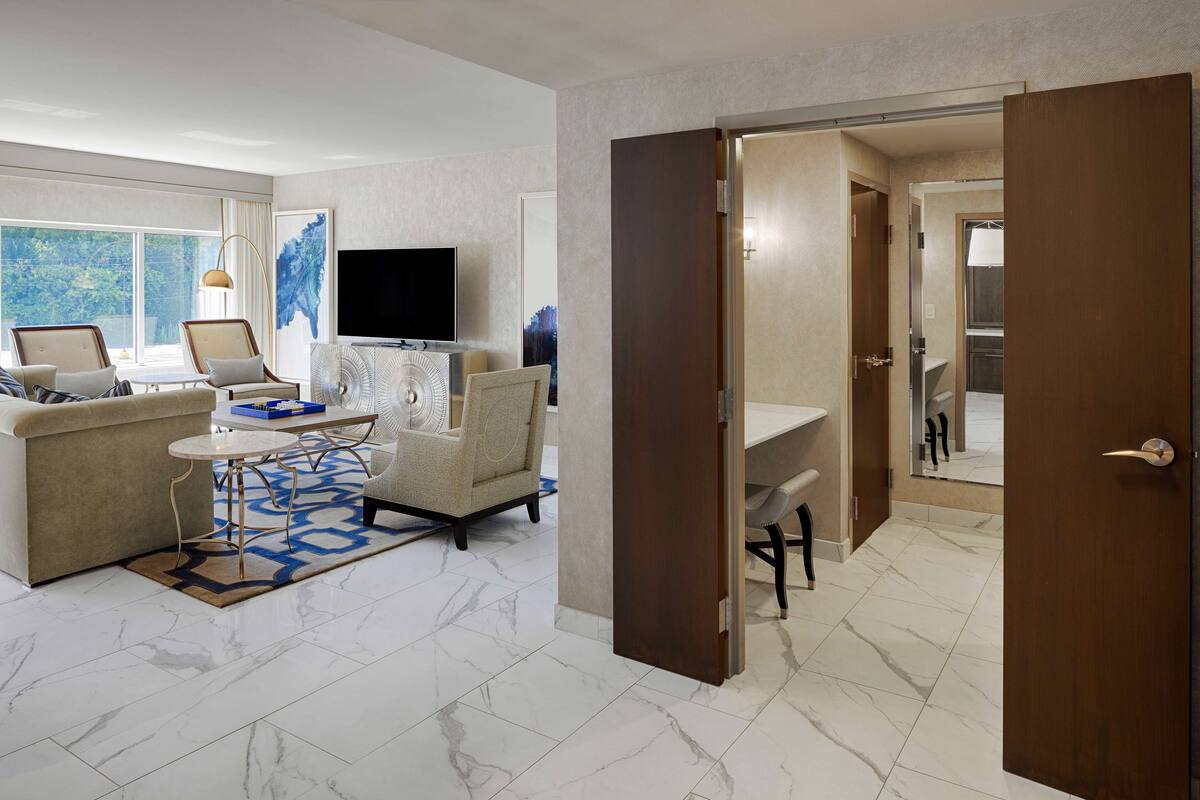 A modern hotel room with a living area featuring a sofa, armchairs, a coffee table, and a flat-screen TV. A large window offers a view of the outdoors.
