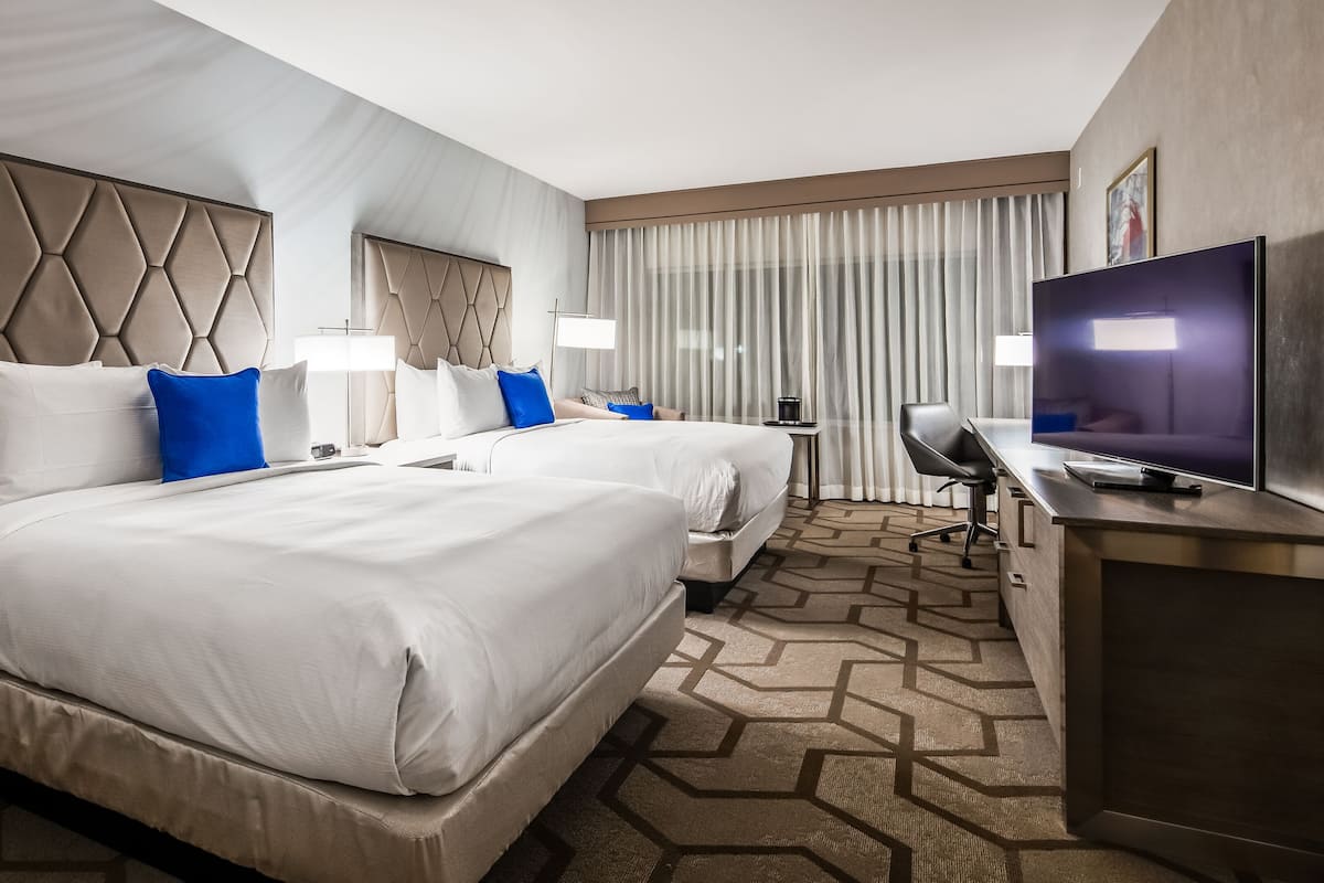 A hotel room with two beds, a TV, a desk, and a window with curtains.